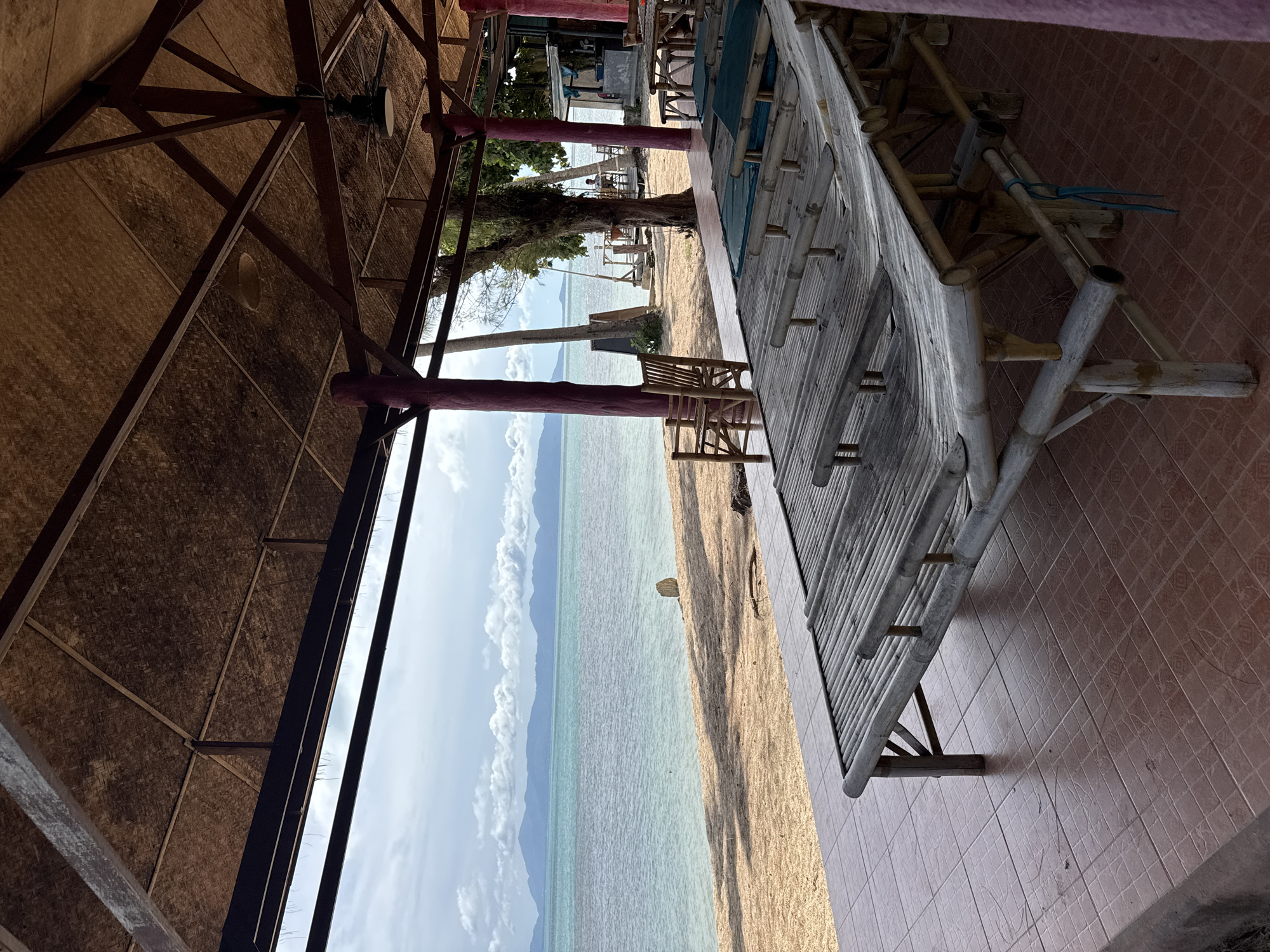 The image depicts a covered outdoor seating area overlooking a beach and the ocean. The structure has a thatched roof supported by wooden beams and purple-painted pillars. In the foreground, there are several low, bamboo daybeds with blue cushions. The floor is tiled in a reddish-brown color. Beyond the seating area is a sandy beach, leading to calm, turquoise water. In the distance, mountains form the horizon under a cloudy sky. There are no people clearly visible in the foreground seating area, but a few individuals can be seen further down the beach, near some buildings and piers. The scene appears to be a relaxed, tropical setting, likely a beachfront restaurant or cafe in Baan Tai, Thailand.