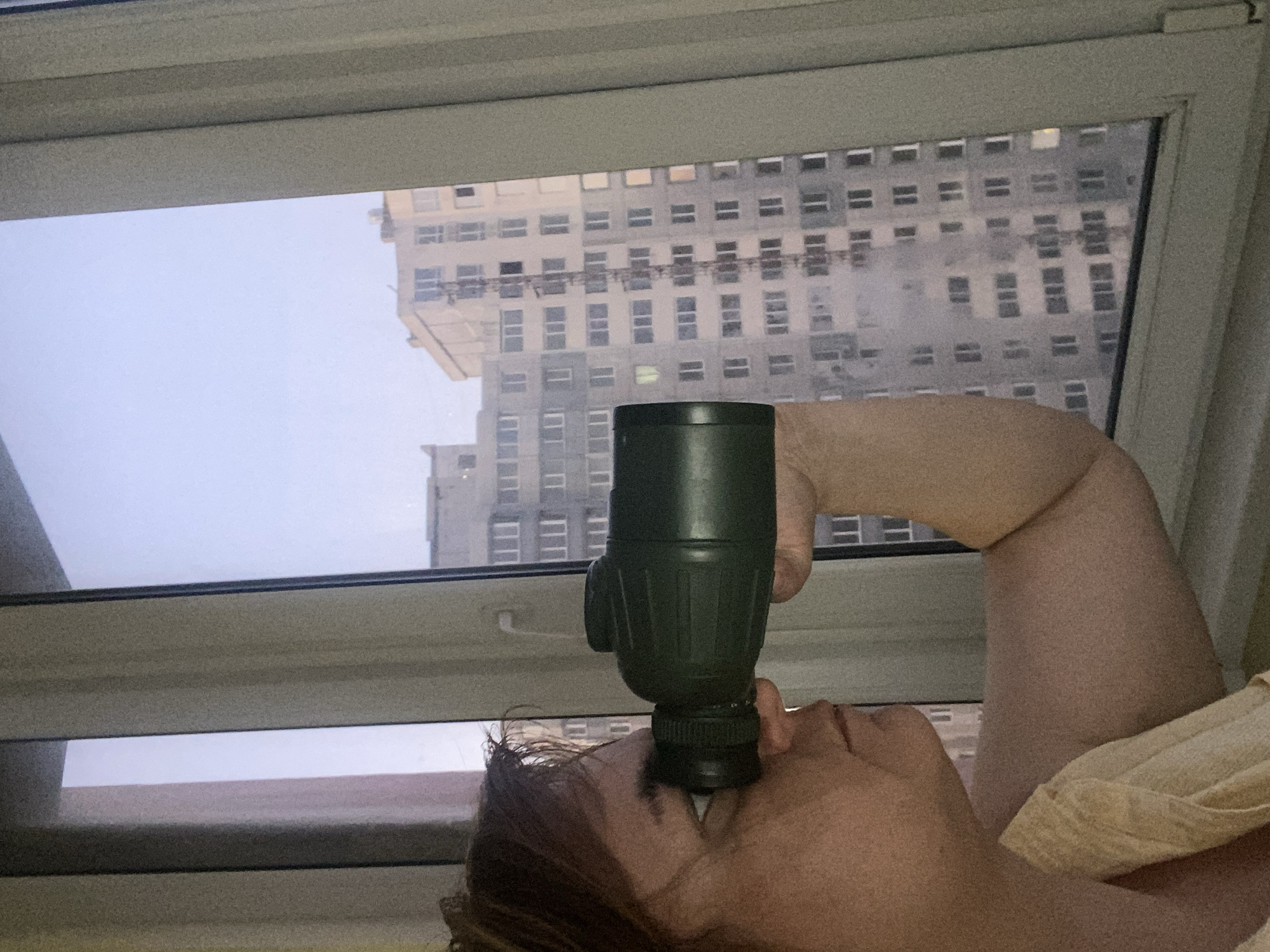 A person is holding a monocular to their eye, looking out a window at apartment buildings in Chornomorsk, Ukraine. The person's face is in profile, with their hair pulled back. The monocular is dark green. The apartment buildings are tall and gray, with many windows. The sky is overcast.