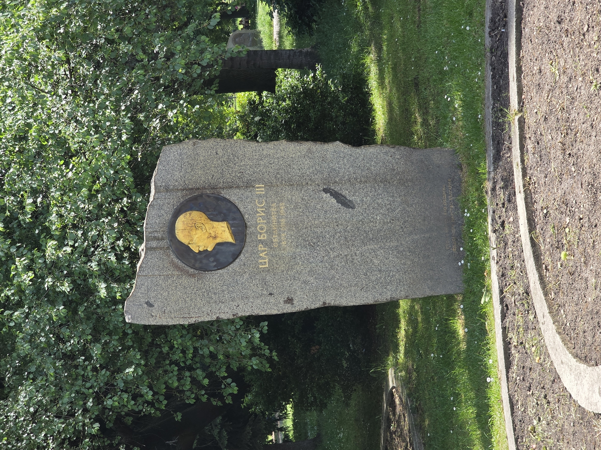 The image shows a gravestone in what appears to be a park or cemetery in Sofia, Bulgaria. The stone is gray and roughly rectangular with a golden profile portrait of Tsar Boris III inset into the upper left corner. Text is inscribed on the stone next to the portrait, identifying the individual and giving dates. The gravestone is positioned in a grassy area bordered by trees and bushes on one side and a concrete edging with soil on the other. Another stone is visible in the background.