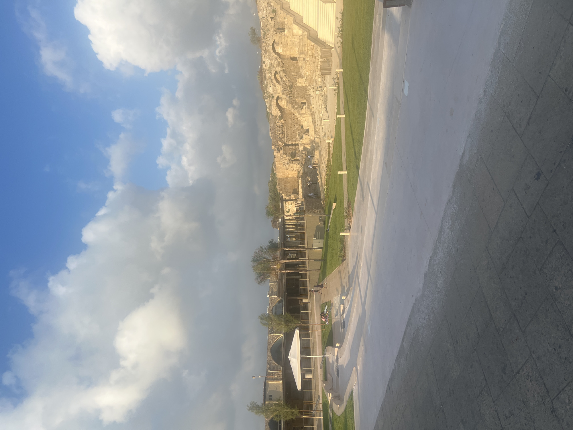 The image shows an archaeological site in Hof HaCarmel Regional Council, Israel, likely Caesarea. In the foreground, a light-colored paved walkway leads into a well-manicured grassy area with some trees and a white umbrella. A group of people are visible in the mid-ground, some sitting on the grass and others walking. Behind them, a long, low-lying building with arched windows and a wooden facade is visible, and further to the right, extensive ancient stone ruins are set against a backdrop of trees and the sky. The sky is blue with scattered white clouds, and the overall lighting suggests it is either morning or late afternoon.