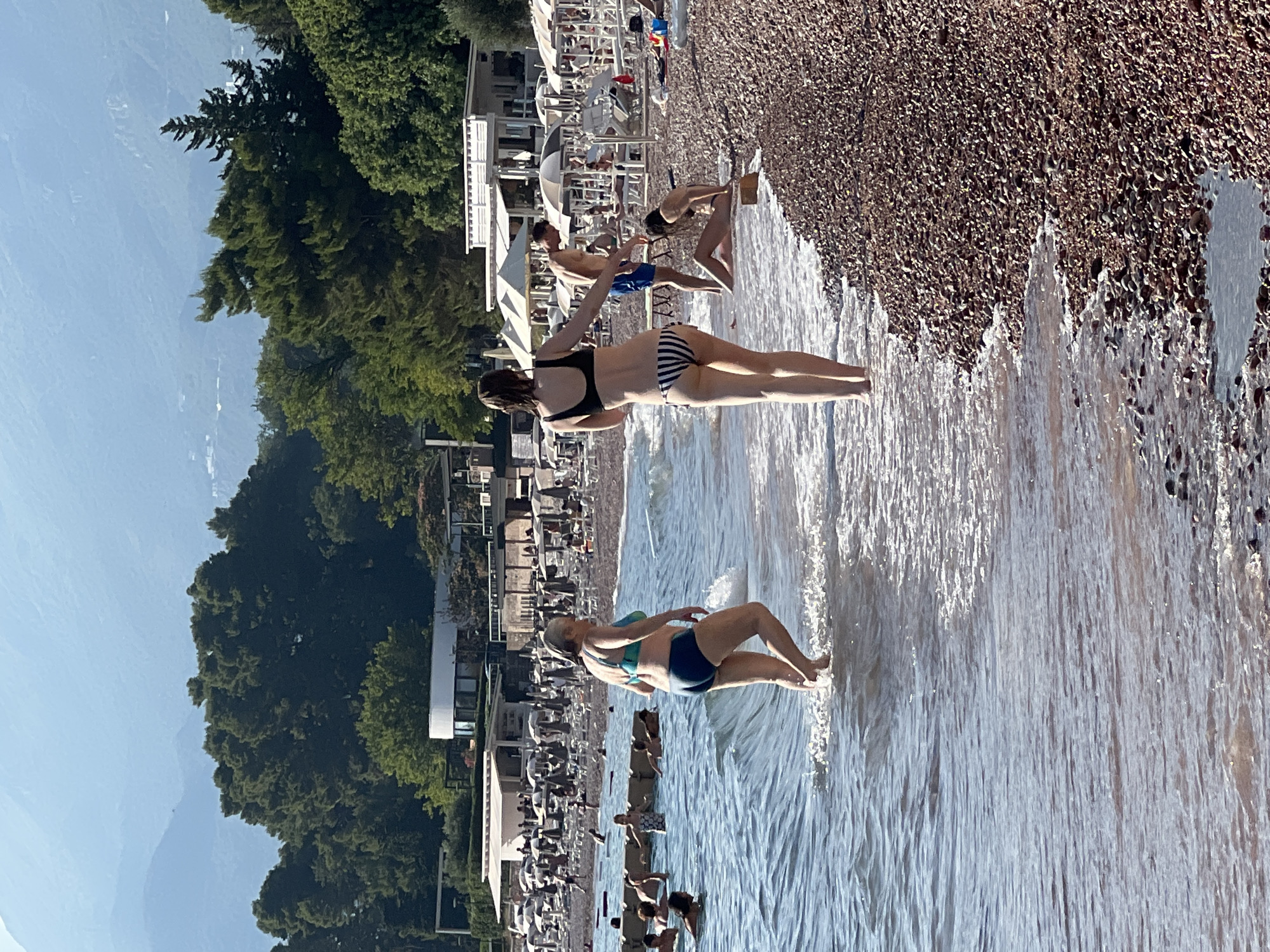 The image depicts a sunny day at a pebble beach in Sveti Stefan, Montenegro. In the foreground, two women are walking along the shoreline. The woman on the left is wearing a teal bikini and the woman on the right is wearing a black and white striped bikini. They are partially submerged in the clear, shallow water. In the middle ground, several other people are enjoying the beach and the water. Some are swimming, while others are relaxing on lounge chairs under umbrellas. The background features a resort area with white buildings and lush green trees, with mountains visible in the distance.