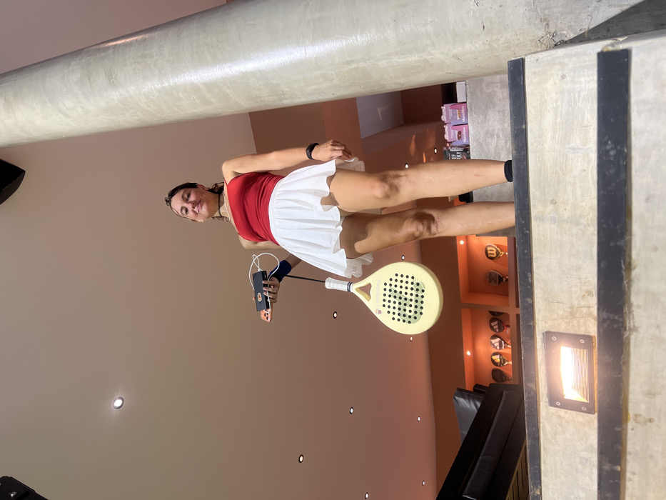 A female subject stands on a concrete staircase in Pecatu, Indonesia, observed from a low-angle perspective. She is dressed in a red tank top and a white pleated skirt. Her left hand holds a light-colored smartphone connected via a white cable to a black portable power bank, and a light yellow padel racket with a perforated circular head is attached to her wrist by a string. A black choker necklace is visible around her neck, and a black smartwatch is on her right wrist. Her expression is a slight smile directed towards the camera. To her right, a large cylindrical concrete pillar extends upwards. The immediate concrete step below her includes an embedded rectangular light fixture. In the background, an indoor area features a high, light orange-colored ceiling with recessed lighting and a mounted black speaker. Further back, a dark sofa is visible on the left, and shelves display various padel rackets and trophies, some branded "Adidas" and "Wilson." Additional shelves contain boxes, some labeled "Munchies." The setting appears to be an interior space, likely a sports club or lounge.