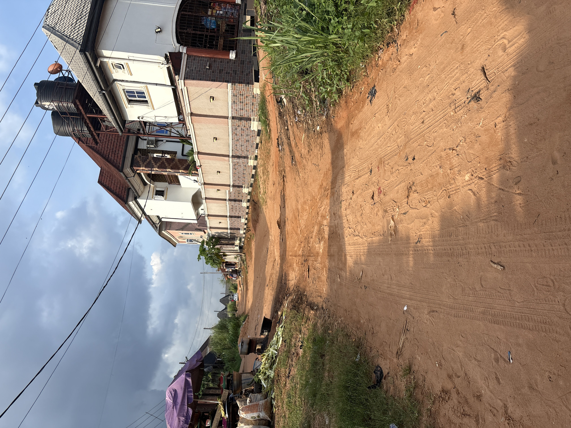 The image depicts a dusty, unpaved road in Obosi, Nigeria. The road is lined with vegetation on the left and modern houses on the right. In the foreground, tire tracks are visible in the reddish-brown dirt. On the left side of the road, there is a market stall with a purple umbrella, sacks of goods, and cooking pots. Further down the road, more houses and some trees are visible. The sky is partly cloudy. A white car is visible in the distance on the road.