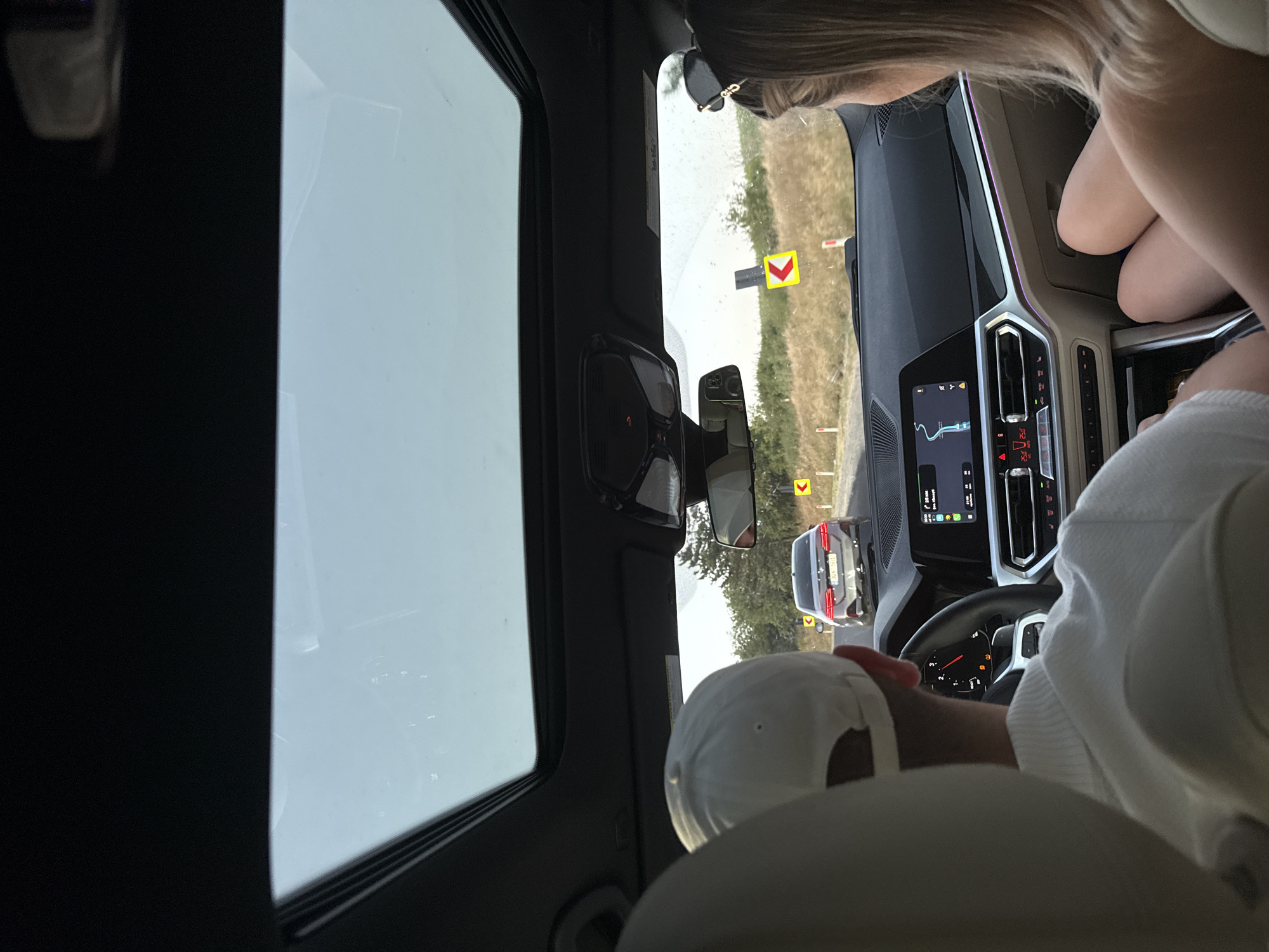 The image, taken from the back seat of a car, shows the interior of a modern vehicle with a panoramic sunroof. In the foreground, the back of a person wearing a white cap and a white collared shirt is visible. To the right, the back of another person's head with blonde hair and sunglasses perched on top is seen. The dashboard of the car displays a navigation screen with a route visible. Outside the car, the road ahead shows a silver sedan driving away. The environment appears to be rural, with trees and dry grass lining the road, and yellow chevron warning signs indicating curves. The sky is bright and overcast. The scene takes place on a road in Fundul Galbenei, Moldova.