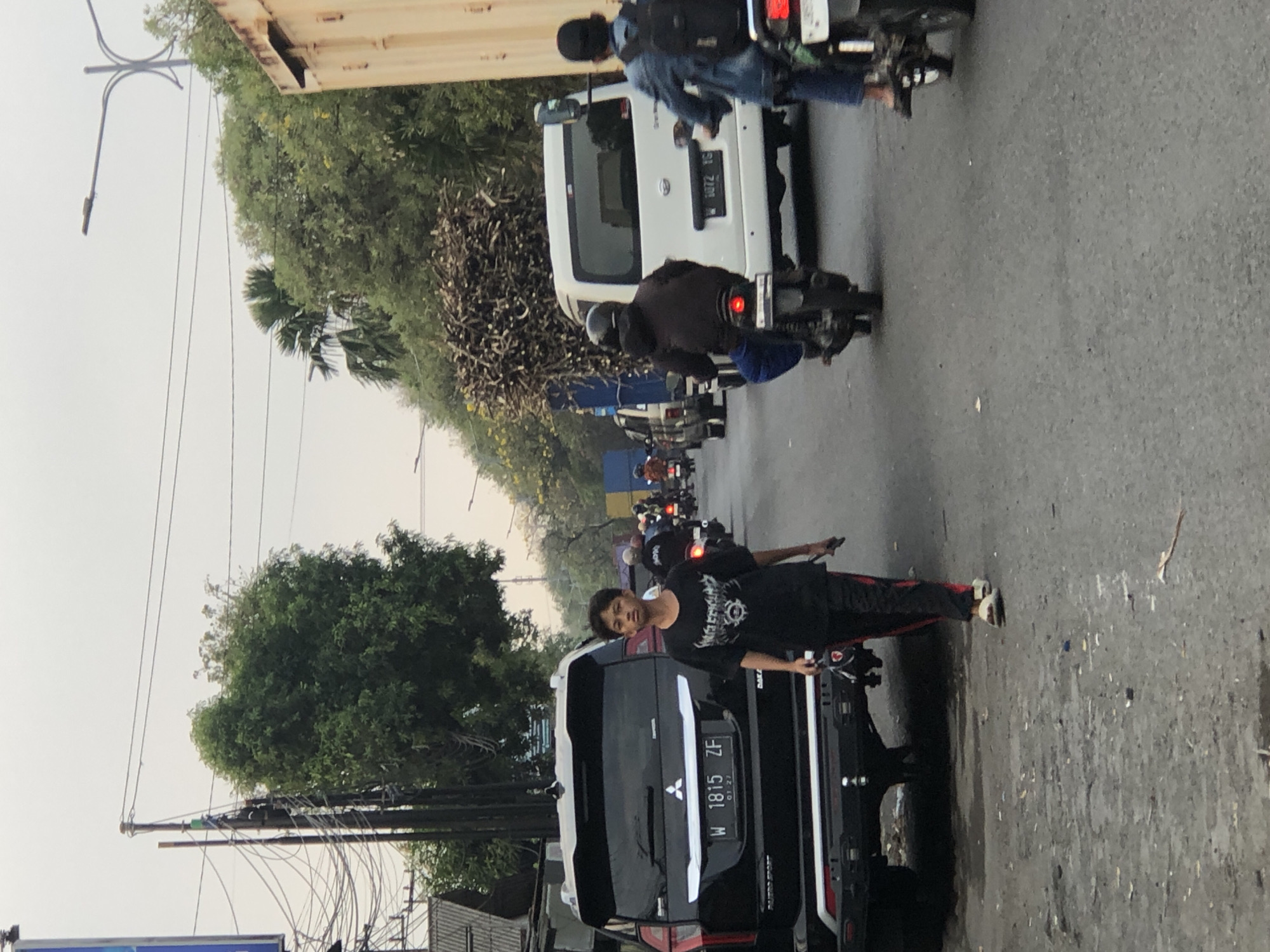 The image depicts a street scene in Ngaban, Indonesia, characterized by moderate traffic and a slightly overcast sky. In the foreground, a young person, appearing to be male, walks from right to left towards the viewer. He is wearing a black t-shirt with a graphic print and black pants with a red stripe, along with white sandals. He is carrying a dark-colored bag or object in his left hand and a dark-colored item, possibly a phone, in his right hand.
To the left of the pedestrian is the rear of a black SUV, identified by a license plate reading "W 1815 ZF". The SUV is parked or moving slowly. Further down the road, traffic consists of motorcycles and a white van. One motorcycle is directly behind the white van, with a rider wearing a dark jacket and blue pants, and a helmet. Another motorcycle is to the far right, with a rider wearing a denim jacket and a backpack. The white van has a license plate that reads "W 1072 IG". A large pile of what appears to be agricultural material, possibly sugarcane stalks, is loaded onto a vehicle or trailer in the background, to the right of the white van. Trees and electrical poles with numerous wires are visible on both sides of the street. The road surface is asphalt and appears somewhat damp and weathered. The overall atmosphere suggests a typical urban or semi-urban traffic environment during daylight hours.