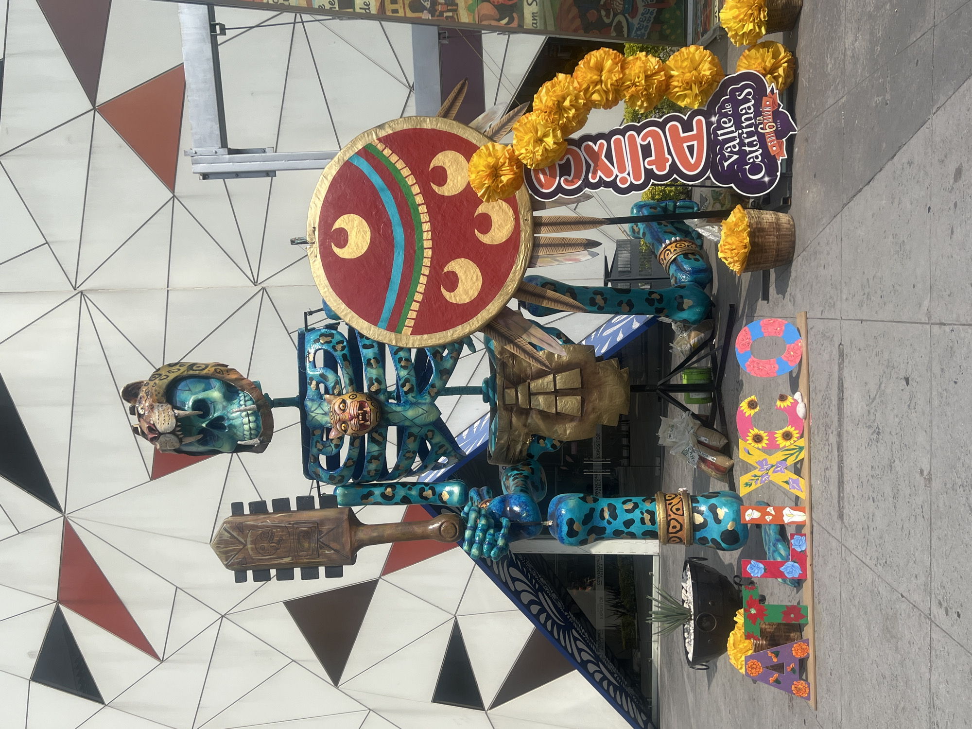 A Day of the Dead themed skeletal figure is seen outdoors in an urban environment. The figure is brightly colored with a jaguar skull and face mask, a red shield with gold lunar symbols and a blue and green band, and a wooden club. The body is patterned with a teal and blue leopard print. A sign reads "Valle de Catrinas Atixco". Yellow marigolds and letters spelling out "MEXICO" are also visible. The background shows a building facade with a geometric pattern of white, brown, and black triangles, and a gray sidewalk.