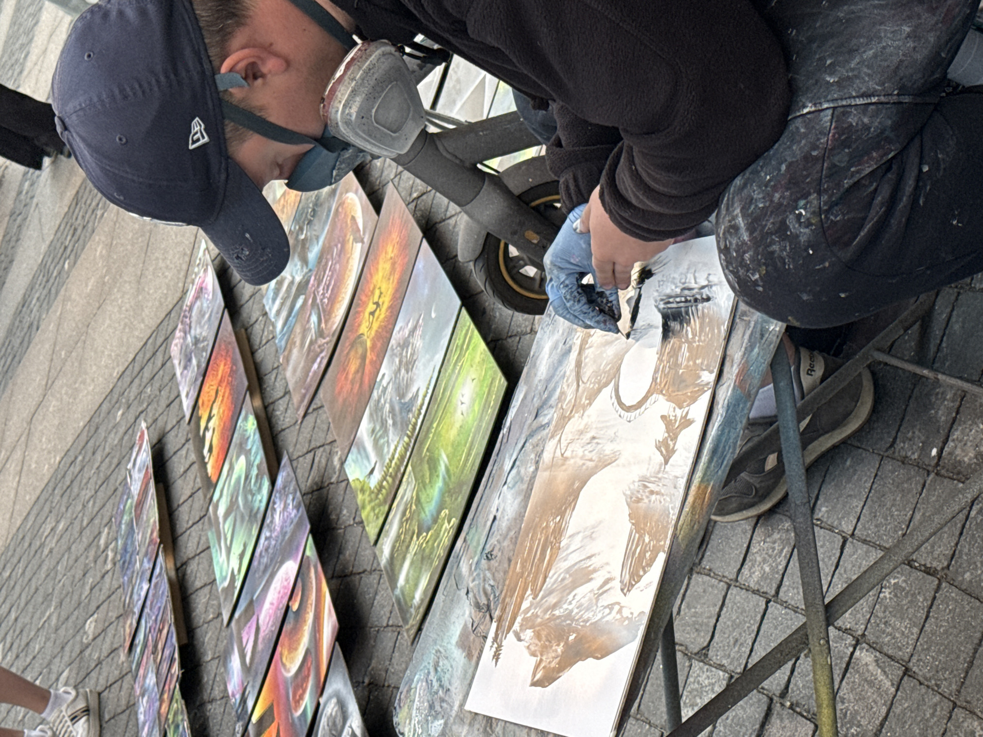 The image shows an artist painting on a street in Saint Petersburg, Russia. The artist is wearing a dark blue baseball cap and a respirator mask, and is dressed in a dark fleece jacket and paint-splattered dark pants. They are wearing light blue gloves and appear to be using an airbrush or spray paint technique. Several completed paintings are displayed on the ground in front of the artist, laid out on wooden planks and a metal stand. These paintings are rectangular and feature vibrant, detailed imagery, including landscapes and fantastical scenes. In the background, other people are visible, and the ground is paved with cobblestones, typical of historic city streets. A portion of a scooter is visible to the right of the artist. The overall scene suggests an outdoor art market or exhibition.