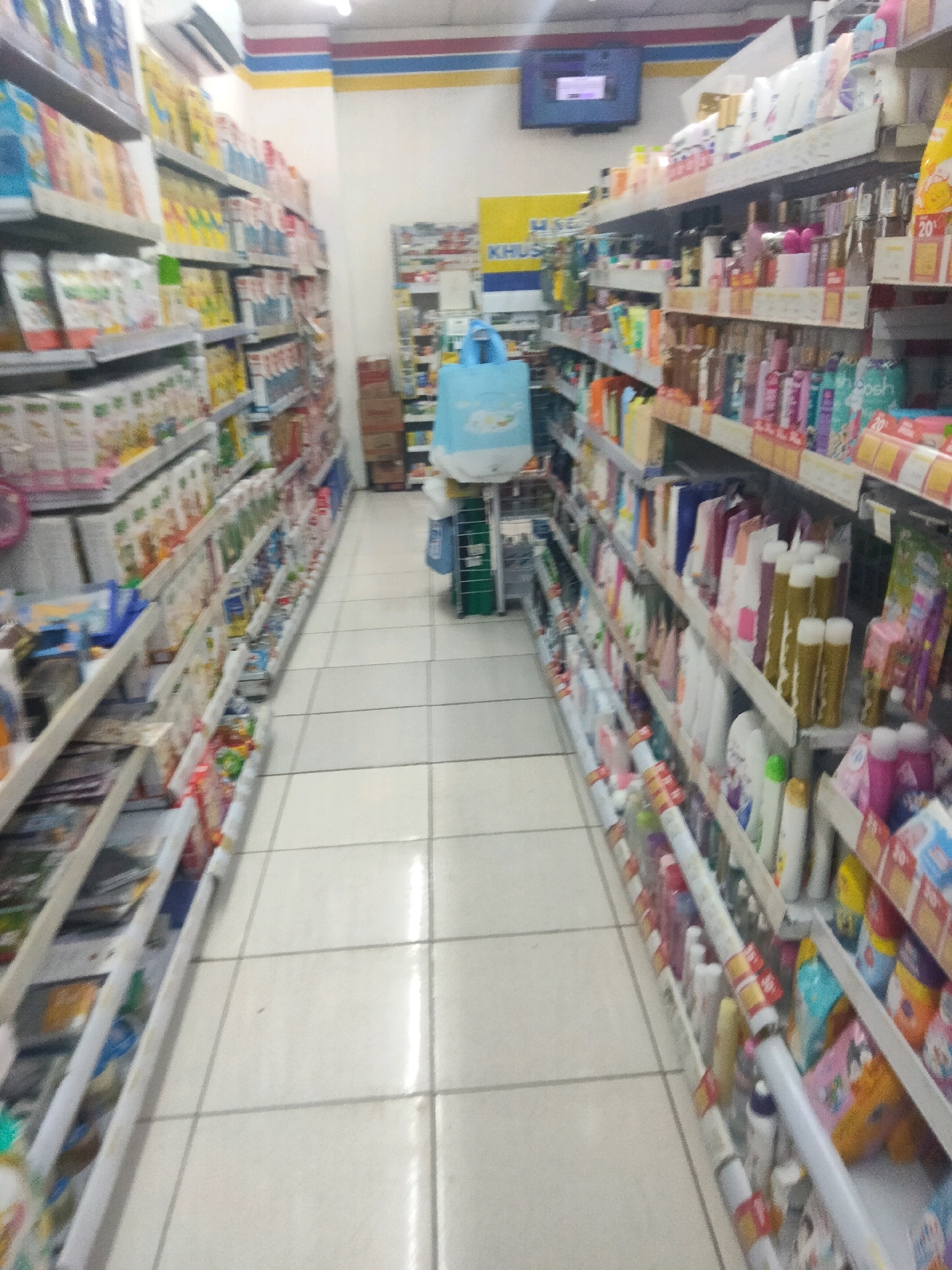 The image depicts an interior view of a convenience store, likely a minimarket, with shelves stocked with various products. The store appears to be located in Pisangan Jaya, Indonesia, as indicated by the context. The perspective is from a low angle, looking down an aisle between two sets of shelves. The shelves on the left are filled with packaged goods, possibly food items or household supplies, exhibiting a range of colors and designs. On the right, the shelves display a variety of personal care products, including bottles, tubes, and boxes in an assortment of colors and sizes. A blue shopping bag is visible on the floor near the front of the right-hand aisle, and a television screen is mounted on the wall in the background. The floor is tiled in a light color. The overall impression is one of a well-stocked retail environment.