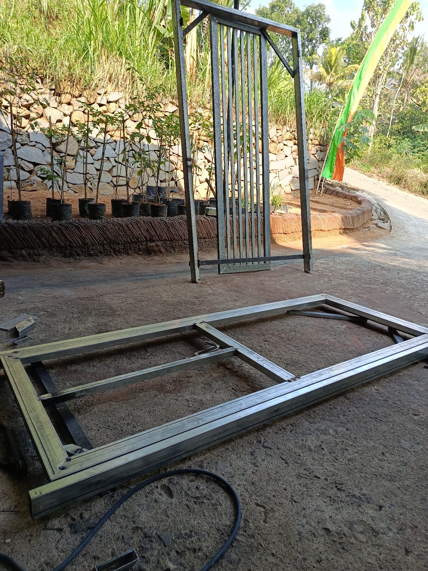 In the city of Lempong, Indonesia, an outdoor scene depicts the construction of a metal gate. In the foreground, a large rectangular metal frame lies on a concrete surface, appearing to be part of the gate's structure. Behind it, a partially assembled gate stands upright, composed of vertical metal bars set within a frame.
The background reveals a tiered hillside with a stone wall, supporting rows of potted plants with slender stems and green leaves. Above the plants, tall green grasses grow abundantly. To the right of the gate structure, a vibrantly colored flag, featuring stripes of green, yellow, and orange, is tied to a pole. The ground consists of concrete in the foreground, transitioning to dirt and a curved, reddish-brown embankment. A winding dirt road is visible in the distance, flanked by lush green vegetation and trees, suggesting a rural or semi-rural environment. The sunlight indicates it is daytime.