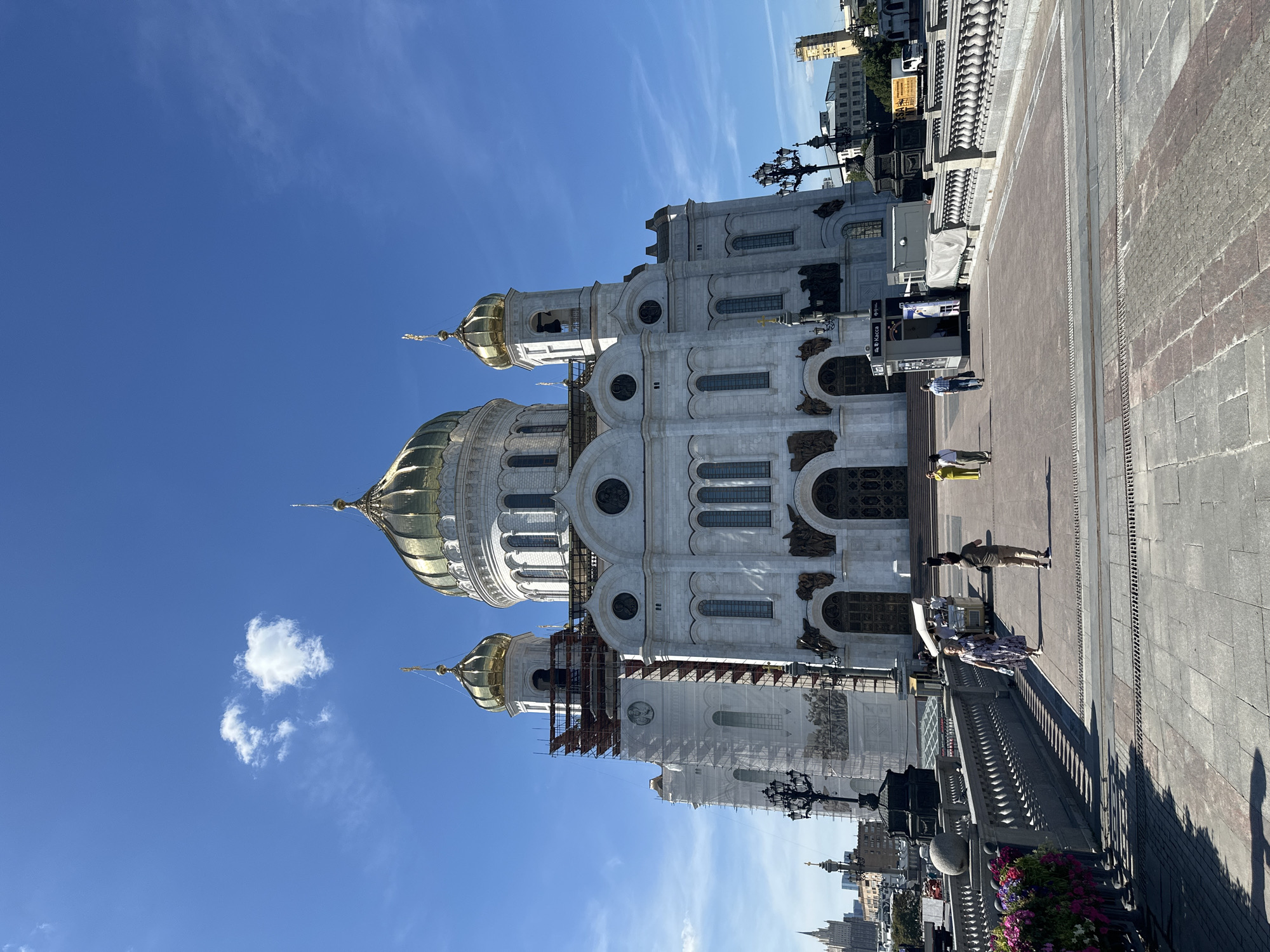 The image captures a wide-angle, eye-level view of the Christ the Savior Cathedral in Moscow, Russia, on a sunny day with a clear blue sky and a few scattered clouds. The grand white cathedral, adorned with multiple golden domes, dominates the upper half of the frame. Scaffolding and white netting are visible on the left side of the cathedral, suggesting ongoing construction or maintenance.
In the foreground, a wide, paved pedestrian bridge leads towards the cathedral. People are seen walking across the bridge and in front of the cathedral's entrance. On the bridge, a woman in a patterned dress walks towards the camera, while a man in a dark t-shirt and light trousers walks away from it. To the left of the bridge, flowering plants in planters add a touch of color.
Further back, to the right of the cathedral's entrance, a small kiosk with the word "Касса" (Kassa, meaning "ticket office") is visible. Beyond the cathedral, a cityscape of Moscow can be seen, with various buildings and structures, including a tall skyscraper in the distance on the left. The overall scene depicts a vibrant public space in the heart of Moscow, showcasing a prominent religious landmark.