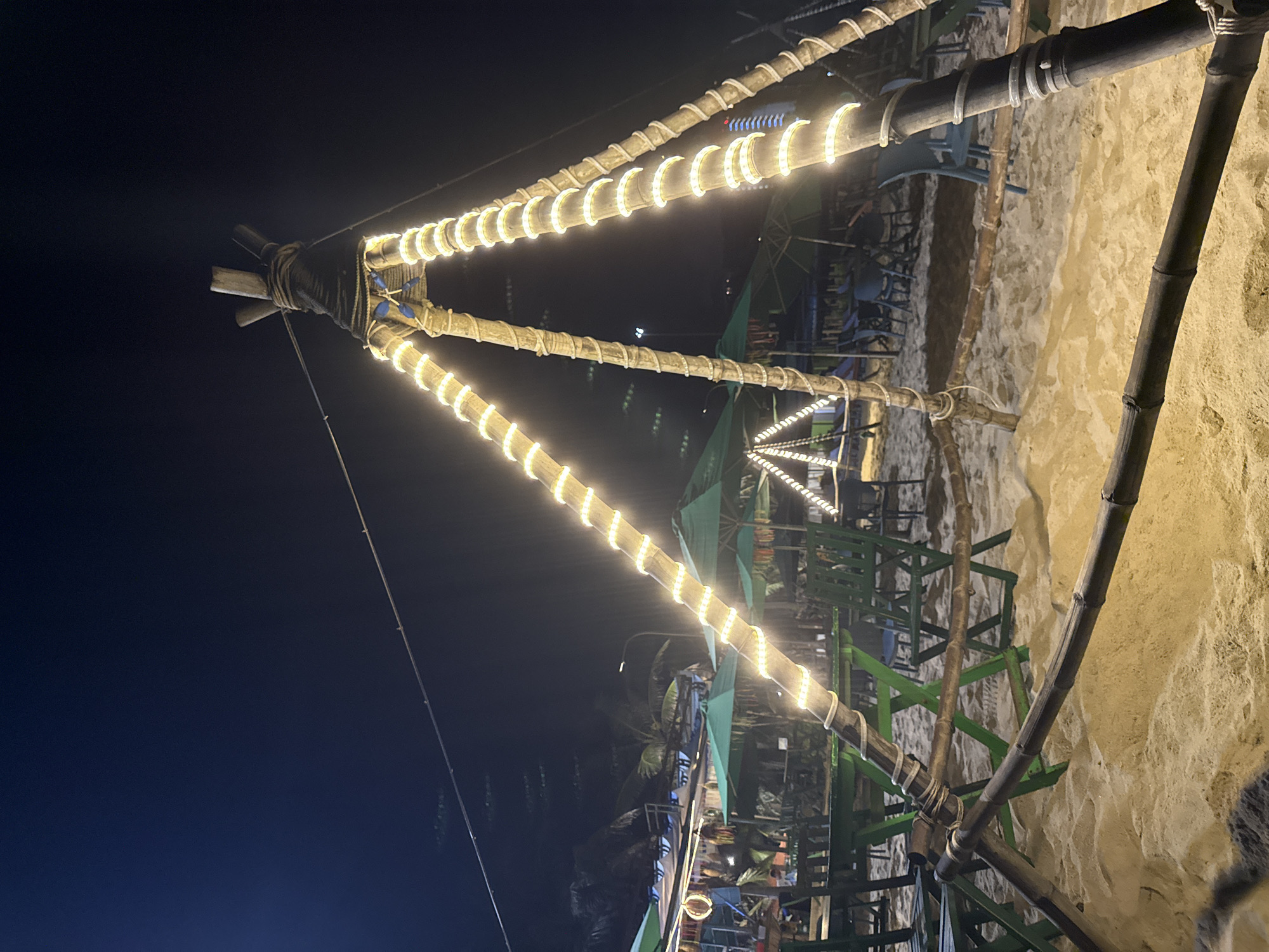 A nighttime shot depicts a triangular structure made of bamboo poles adorned with strings of lights. The structure is set against a dark blue sky on one side and a sandy area with vegetation on the other. Green umbrellas and tables and chairs are visible in the background, hinting at an outdoor dining or gathering space. The lights outline the triangular shape of the bamboo frame, creating a warm glow. The location is likely in Đà Nẵng, Vietnam, based on the style of construction and the surrounding environment.