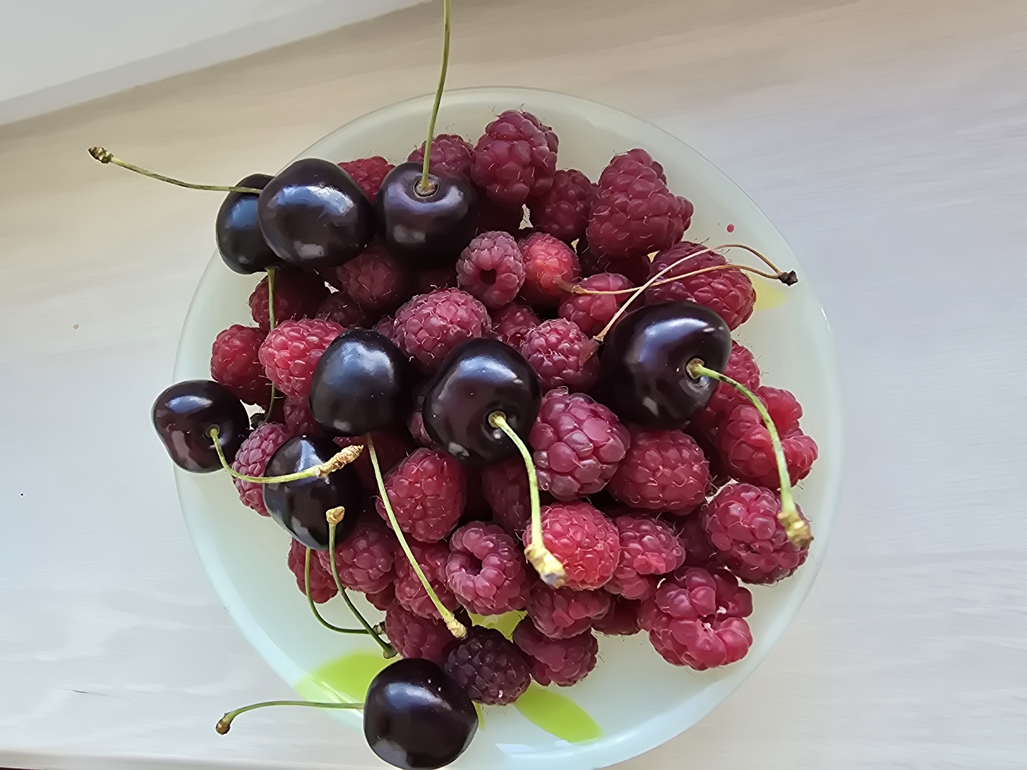 A close-up, high-angle shot shows a light green, circular plate filled with fresh raspberries and dark cherries. The plate rests on a white, textured surface, likely a table. The berries are piled high, with some cherries nestled amongst the raspberries, and others perched on top with their stems intact. The raspberries are a vibrant pink-red, while the cherries are a deep, glossy dark red. The lighting is bright, casting soft shadows and highlighting the plumpness of the fruit. The background is a plain, light-colored wall, suggesting an indoor setting. The image composition is simple and focused on the fruit, implying a still life arrangement. There are no people or animals visible in the scene. The image was taken on July 15, 2023, at 10:26 AM in Sofiivska Borshchahivka, Ukraine.