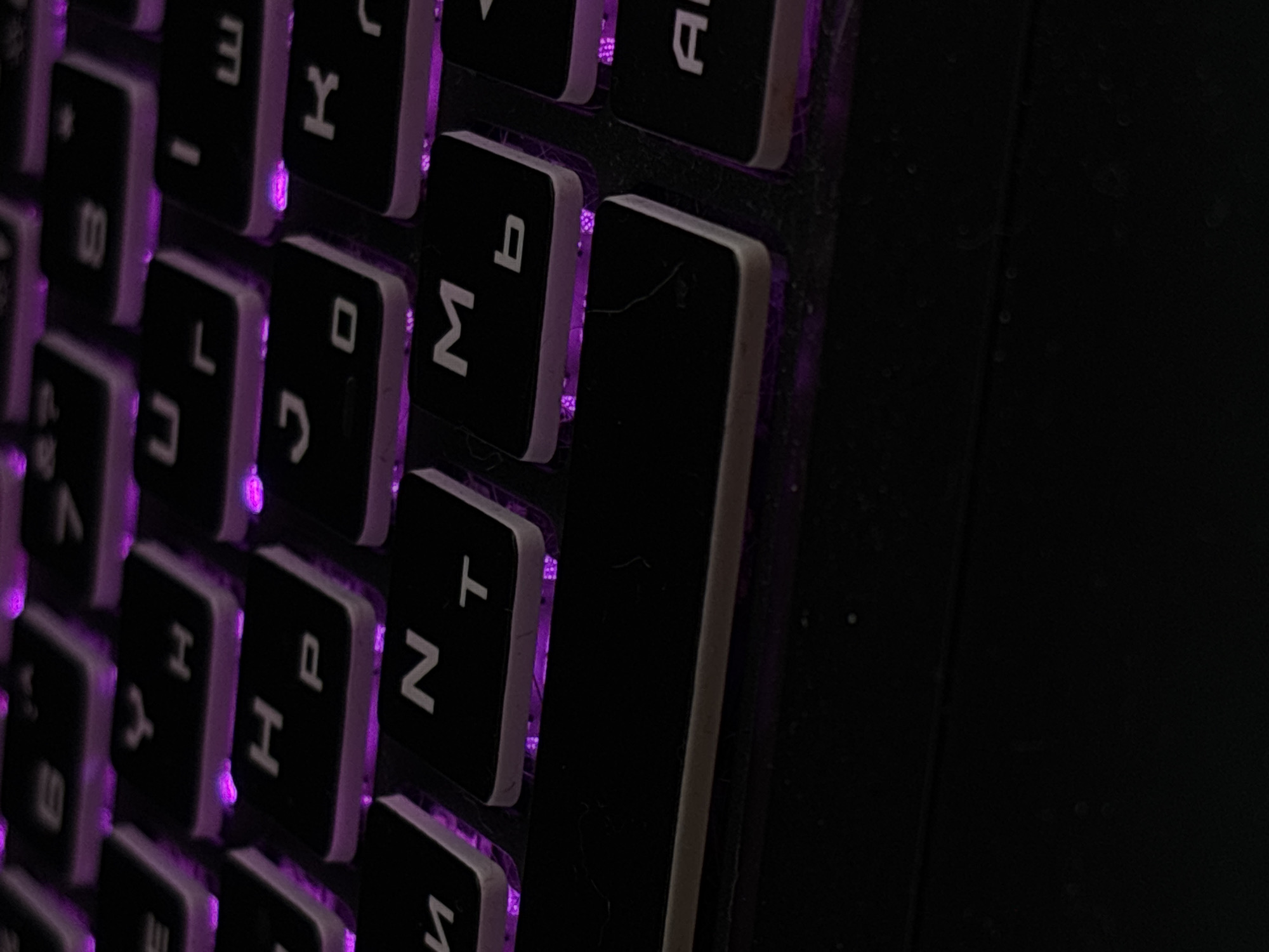 The image shows a close-up of a backlit keyboard with purple lighting. The keys are black with white lettering. Some of the visible keys include "H", "P", "N", "T", "M", "b", "U", "L", "J", "O", and "E". The keyboard appears to be part of a laptop or a gaming setup, given the vibrant backlighting. There are no people or any other distinguishing features in the image. The background is dark, suggesting the photo was taken in low light conditions. The image does not provide any information about its location in Saint Petersburg, Russia, or any other city.