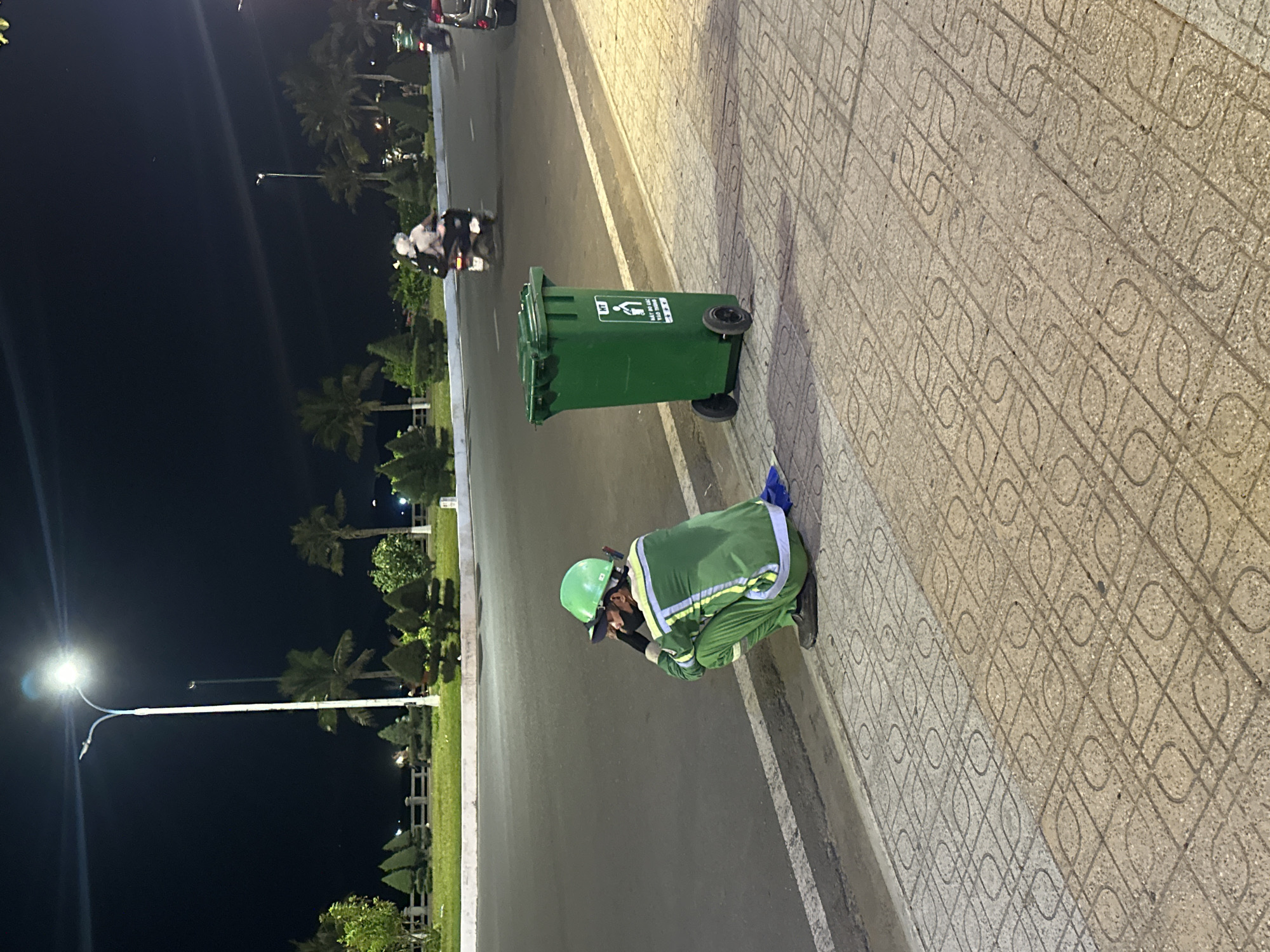 A sanitation worker wearing a green uniform and helmet is crouching near a green trash bin on a sidewalk next to a street in Bac Nha Trang Ward, Vietnam. The street is lit by streetlights. A motorcycle and car are visible in the distance. The sidewalk has a patterned design. The environment includes palm trees and other greenery. The time of day appears to be evening or night.