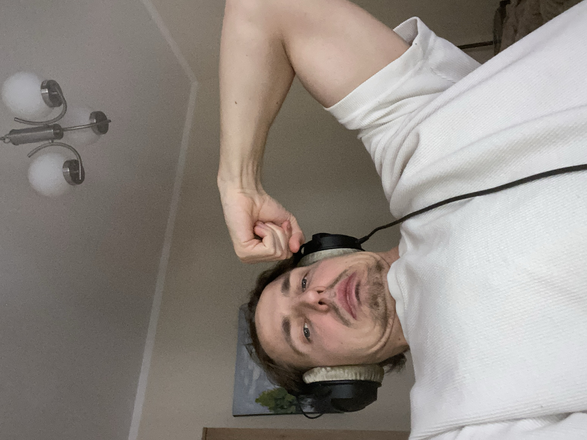 The image displays an indoor scene featuring a male individual, an adult with brown hair, a mustache, and a goatee. He is wearing a white waffle-knit long-sleeved shirt and black over-ear headphones with light-colored earcups. His right arm is flexed, with the hand forming a fist above his head, showcasing the biceps. The man is looking directly at the camera with a strained facial expression characterized by pursed, downturned lips, raised cheeks, and a slight furrowing of the brow. The background consists of a light-colored wall and ceiling. A three-arm chandelier with white spherical light shades is suspended from the ceiling in the upper right quadrant. A small framed artwork depicting green trees and a light sky is visible on the wall behind the man's left shoulder. A dark vertical structural element is partially visible on the far left. The specific location within the city of Šušanj, Montenegro, is not discernible from the visual information provided within the image.