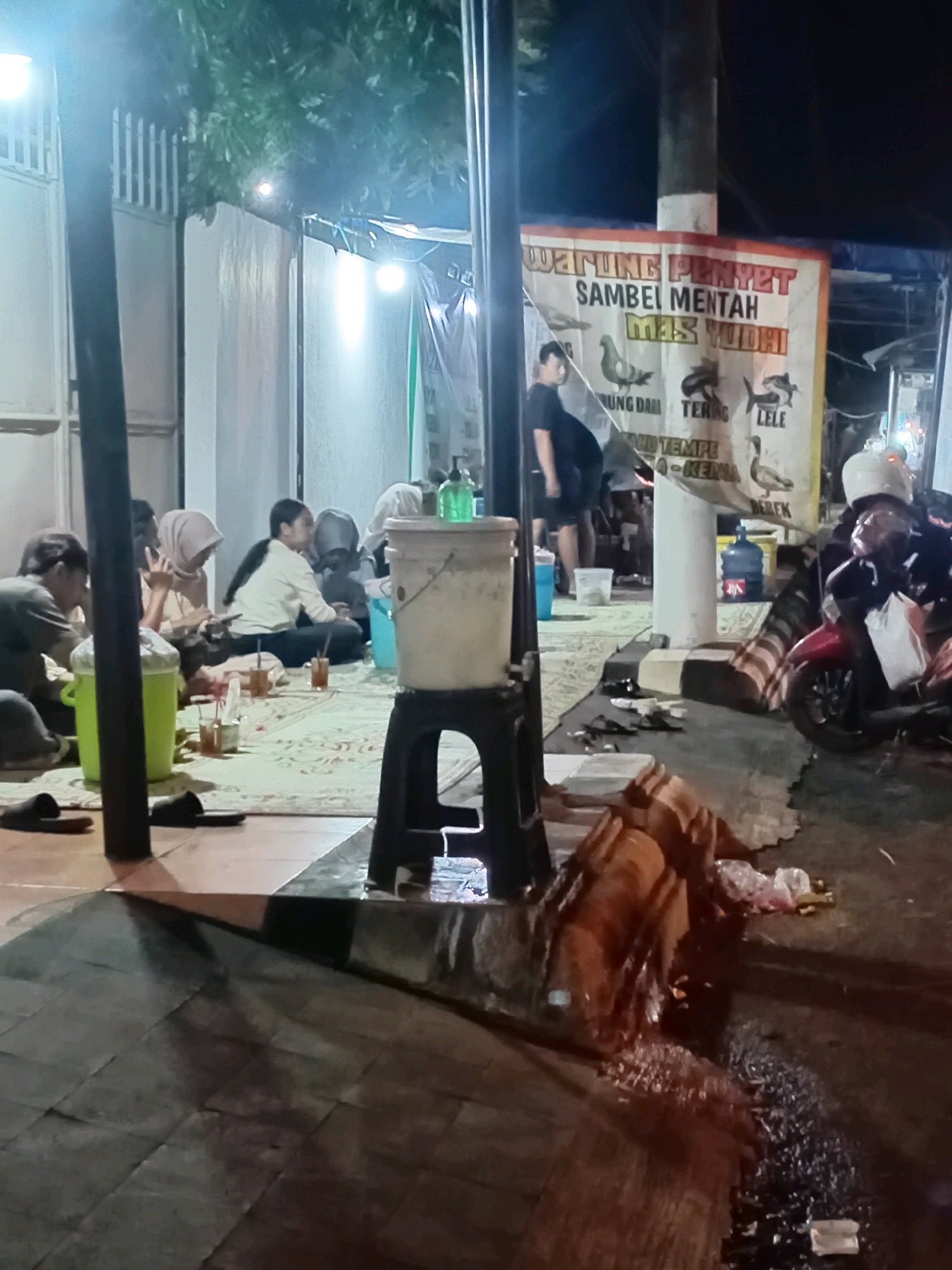 In the city of Deman, Indonesia, a group of people are gathered outdoors at night, likely at a food stall or warung. The scene is illuminated by artificial lighting, creating a slightly hazy and dimly lit atmosphere. Several individuals, some wearing hijabs, are seated on the ground on a patterned mat, appearing to be eating or socializing. In the background, a banner with Indonesian text and illustrations of various animals, including fish, birds, and a duck, is visible. A man in a black t-shirt stands near the banner. To the right, a motorcycle is parked, and a person wearing a helmet is seated on it. Various items like buckets, cups, and bottles are scattered around the area, indicating a casual outdoor dining or gathering setup. The ground is a mix of tiled and paved surfaces.