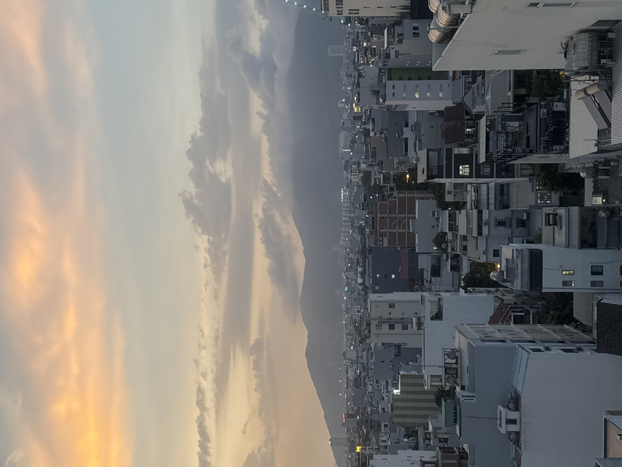 The image shows a dusk view of Da Nang, Vietnam, facing west towards the Annamite Mountain Range. The sky is partially clouded, with orange and gray hues. A dense urban landscape with buildings of varying heights stretches from the foreground toward the mountains. Lights are visible in many buildings, suggesting early evening. The buildings appear predominantly residential, with a mix of architectural styles. No people are visible.