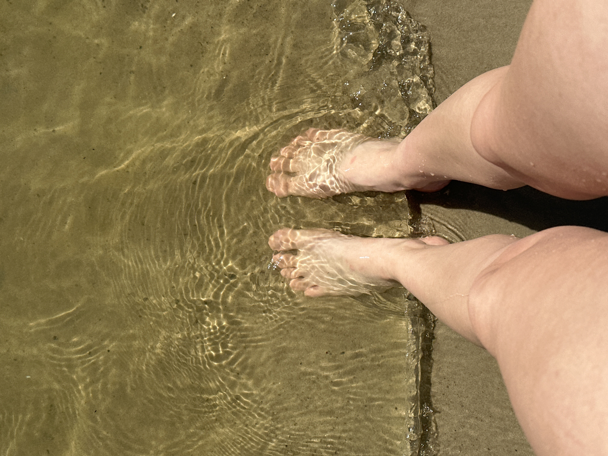 The media file displays a close-up, top-down view of a person's bare feet and lower legs partially submerged in shallow, clear water. The water's surface is rippled, creating a pattern of light and shadow on the sandy or muddy bottom beneath. The feet are positioned in the water, with visible toes and the tops of the feet. The skin appears fair, and the water is casting dappled light onto it.
The scene is set at the edge of a body of water, likely a beach or shoreline, as evidenced by the sandy texture visible at the top right of the image. The lighting suggests it is daytime, with the sun's rays creating the characteristic patterns on the water's surface. The overall impression is one of relaxation and a simple enjoyment of being in or near water. There is no visible text in the media file. The location context provided is Horishni Plavni, Ukraine.