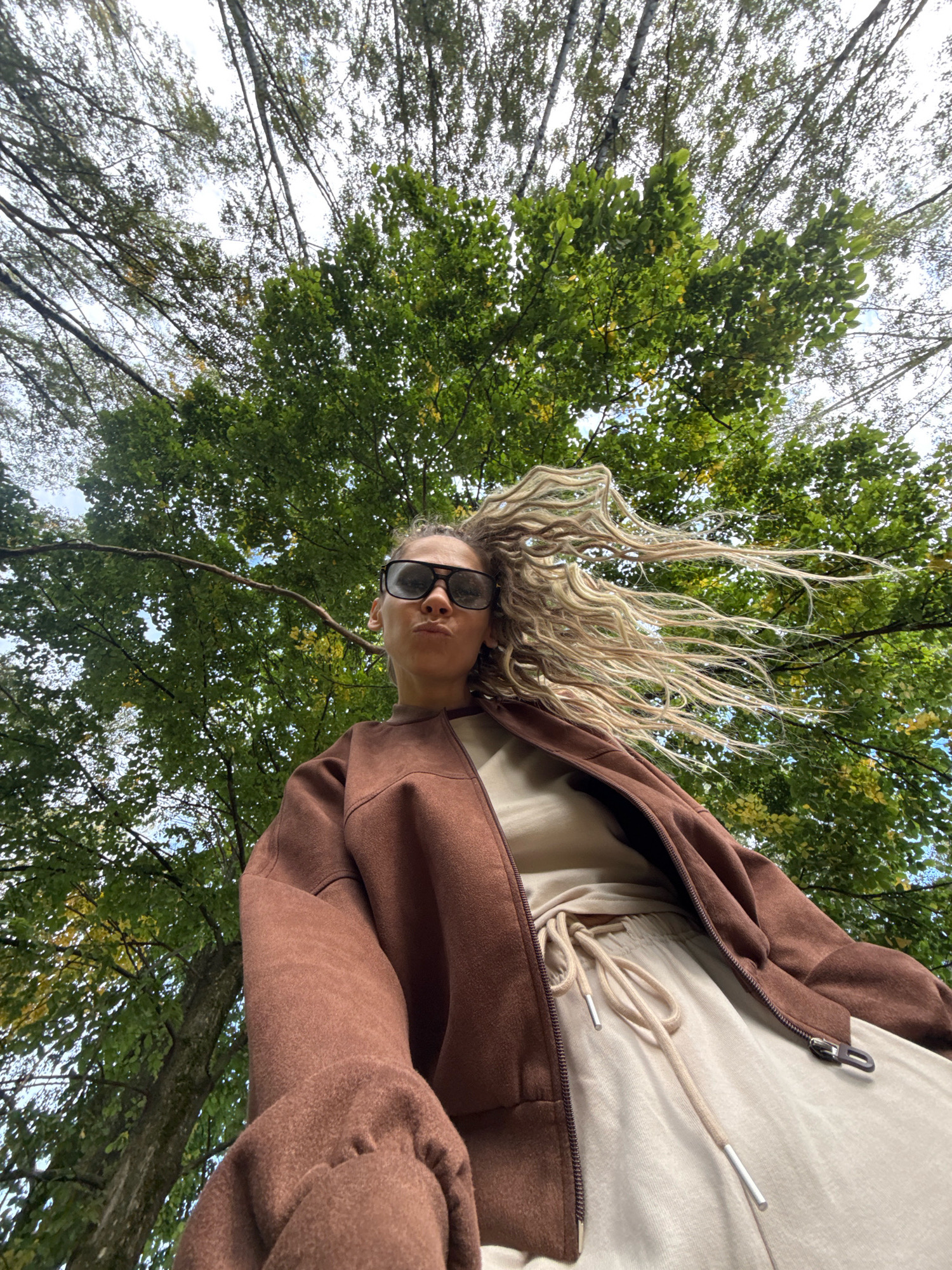 A low-angle, Dutch-tilted shot captures a woman from the chest up, standing outdoors. She is wearing a brown zip-up jacket over a light-colored t-shirt and light-colored sweatpants. The woman has long, blonde, wavy hair styled into what appears to be dreadlocks, which are blowing to her right. She is wearing dark sunglasses and her lips are pursed. The background is dominated by lush green trees and a bright sky, suggesting an outdoor, possibly park or forest, setting. The image is sharp and well-lit. The location is specified as Rusanovo, Russia.