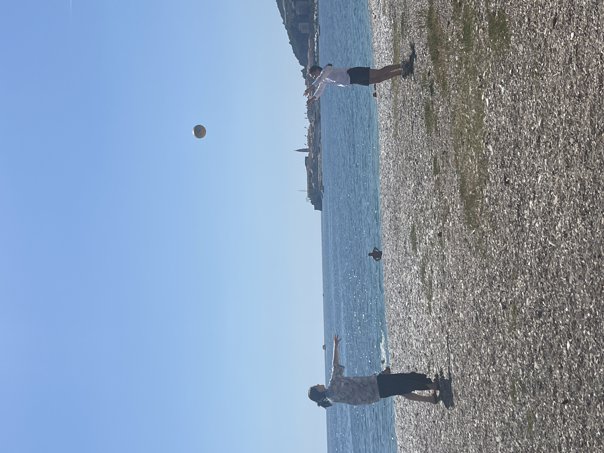 The image shows two people playing with a ball on a pebble beach. The scene is set in Budva, Montenegro, indicated by the coastline and distant buildings. The sky is clear blue with the ball in mid-air. One person, facing away from the camera, wears a light shirt and dark shorts with arms outstretched. The other person, in the foreground, is also positioned with arms raised toward the ball and wears a patterned shirt and dark shorts. The sea stretches between them, sparkling in the sunlight.