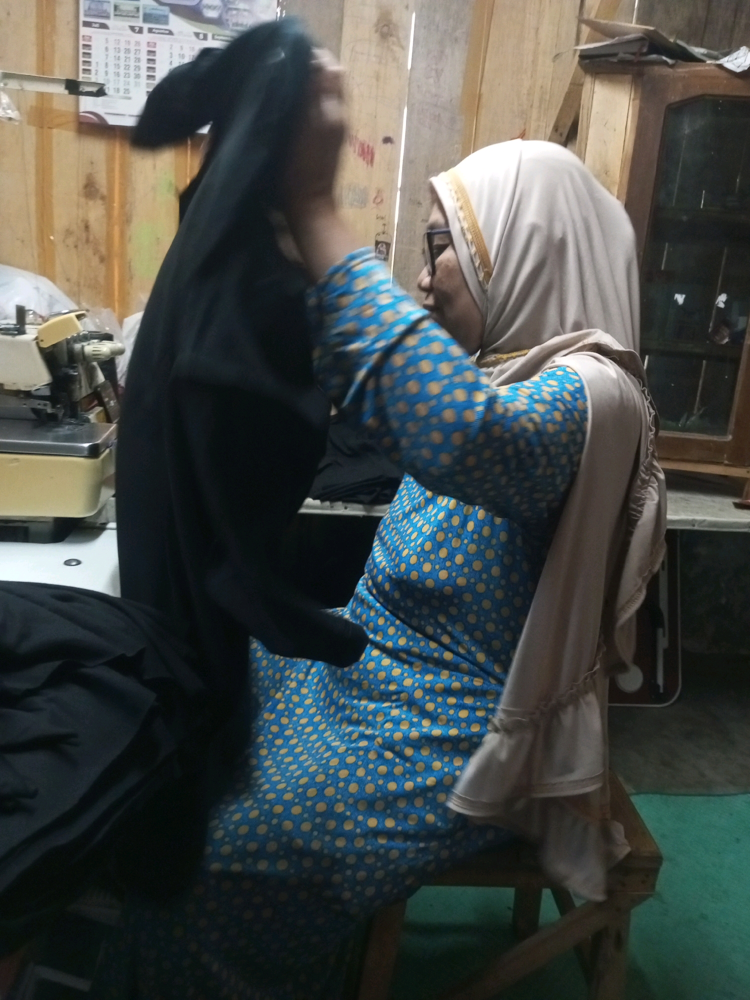 In Kalibeber, Indonesia, a woman wearing a beige hijab and glasses is in a room with wooden walls. She is wearing a blue dress with yellow polka dots and a beige shawl with ruffled sleeves. In front of her, a black garment, possibly a burqa or abaya, is being held up, obscuring her face. To her left, a sewing machine is visible on a table. The floor is covered with a green carpet.