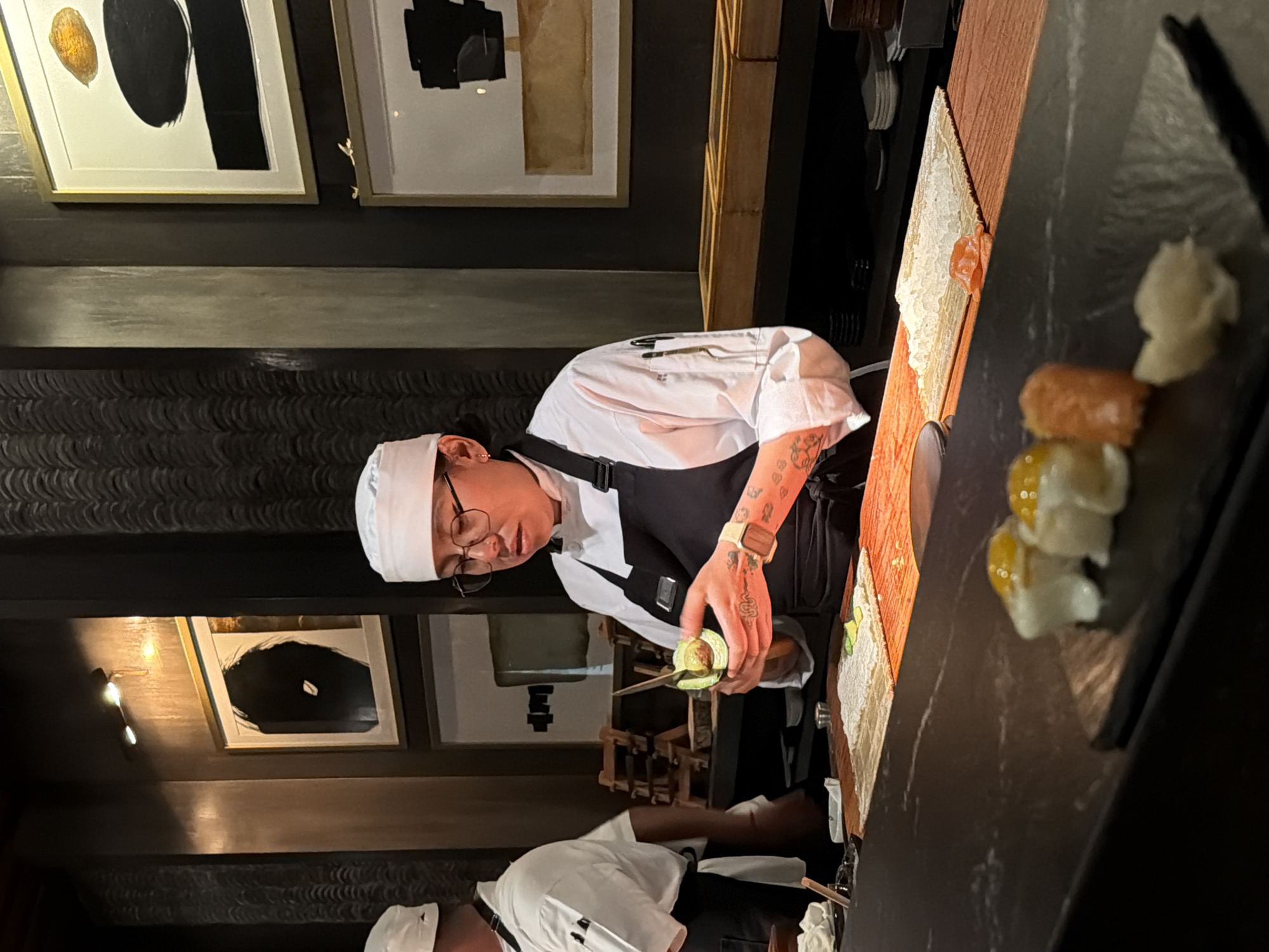 A chef in Puebla, Mexico is preparing sushi. The chef, wearing a white uniform and glasses, is holding a tool and working over a counter. Sushi pieces are visible on a black plate to the right. Behind the chef, there are abstract art pieces hanging on the wall. Another person in a white uniform is seen slightly behind and below the main chef.