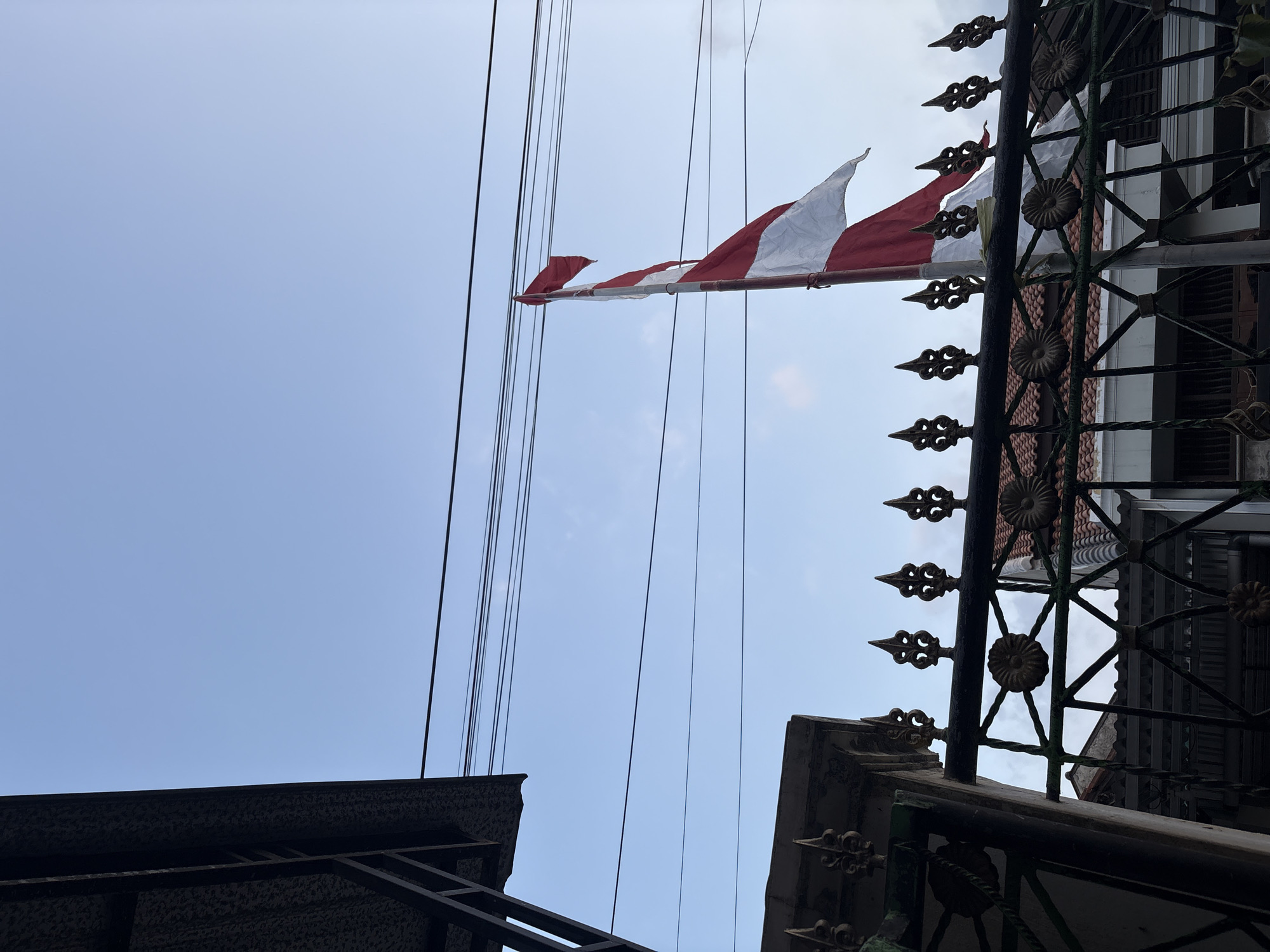 The image captures a partial view of a red and white flag, likely the Indonesian flag, flying on a flagpole. The flag is partially unfurled and appears to be made of fabric. It is positioned against a pale blue sky with some scattered clouds. In the foreground, a dark, ornate metal fence with decorative elements is visible. Behind the fence, rooftops of buildings can be seen, suggesting an urban or semi-urban environment. Several electrical wires run horizontally across the sky. The overall lighting suggests it is either early morning or late afternoon. The location is identified as Palimanan, Indonesia.