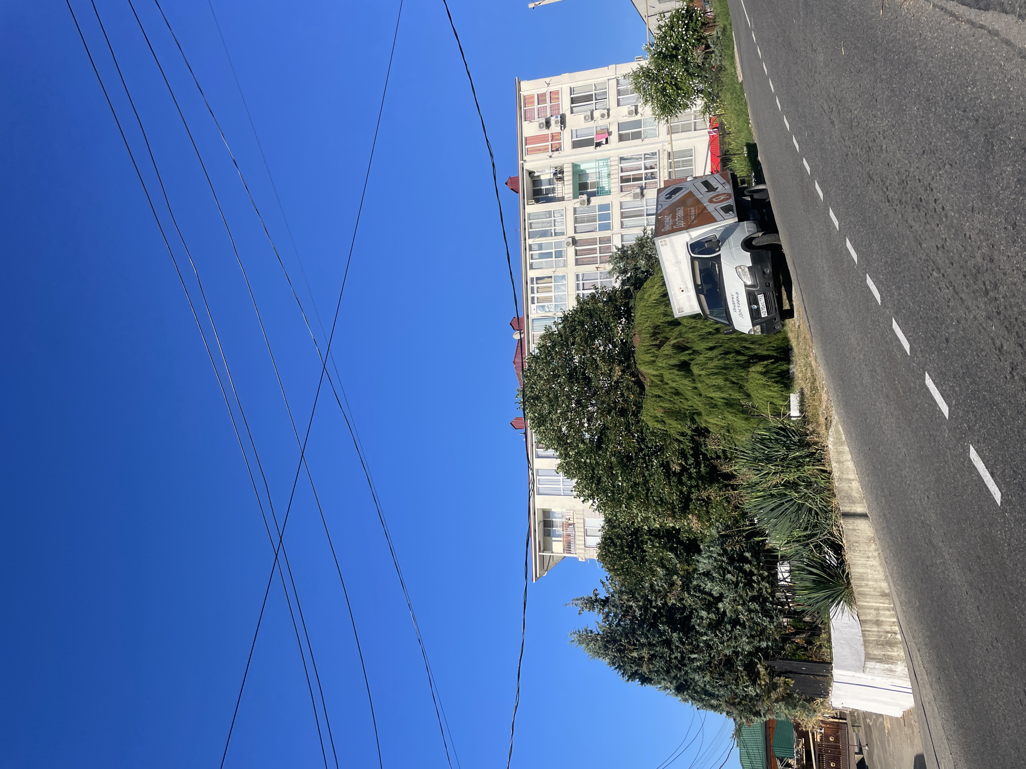 The image shows a street scene in Baranovka, Russia, on a clear day. A white delivery truck with "Yandex Delivery" branding is parked on the right side of the uphill road, angled towards the left. The truck has a license plate that reads "H270YP 197". To the right of the truck, a multi-story white apartment building with numerous windows and balconies is visible. In front of the building and to the left of the truck, a variety of trees and bushes are planted, including coniferous trees and agave plants. The road is paved with asphalt and has white dashed lane markings. Overhead, several electrical wires crisscross the clear blue sky.