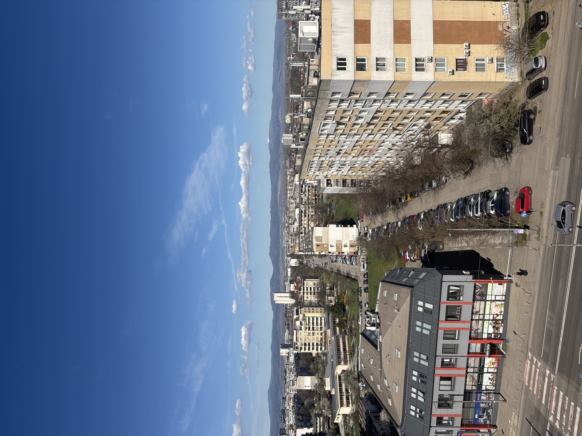 The image presents an elevated view of a cityscape, possibly taken from a high-rise building. The sky to the left is a vibrant blue with scattered white clouds. A mountain range stretches along the horizon. The urban area is characterized by a mix of building types, including large apartment blocks and smaller structures. Rows of parked cars line the streets, and other vehicles are visible in motion. The location is suggested to be Sofia, Bulgaria, based on the architecture and urban layout. The focus is on the buildings, streets, and a few trees, with no people clearly identifiable.