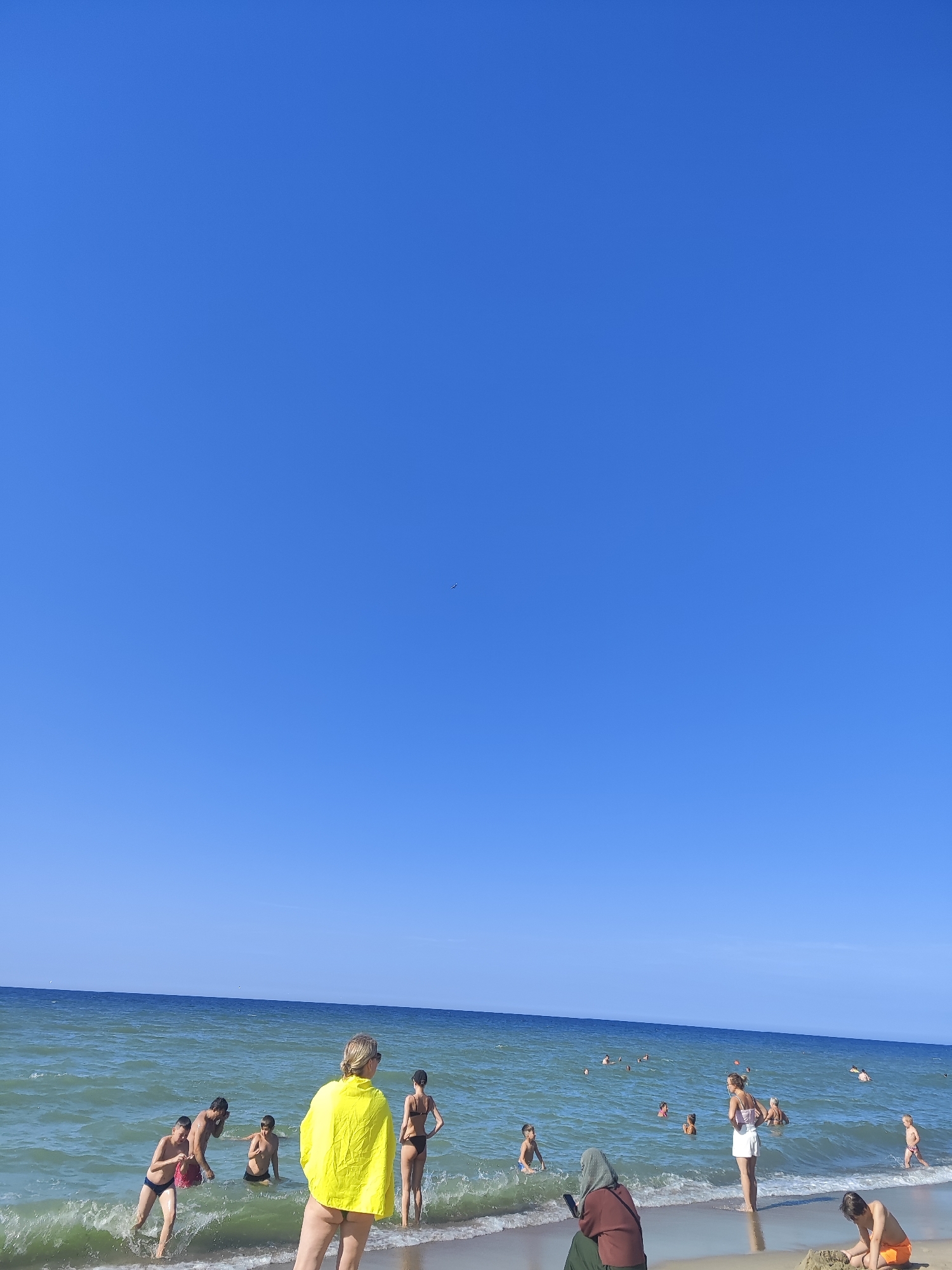 A clear, sunny day at the beach in Zelenogradsk, Russia, with a bright blue sky dominating the upper portion of the image. The Baltic Sea is calm with gentle waves lapping the shore. A group of people are scattered along the beach and in the water. In the foreground, a woman in a yellow towel is facing away from the camera, looking out at the sea. To her left, a man and two boys are wading in the water, with one boy holding a pink bag. To the right of the woman, another woman in a bikini stands on the shore. Further out in the water, several other individuals are swimming or playing. On the right side of the frame, a young boy in orange swim trunks is building a sandcastle. A woman in a maroon hijab and green skirt is seated on the sand, looking at her phone. The beach is sandy, with a few patches of dry sand and some wet sand near the water's edge.
