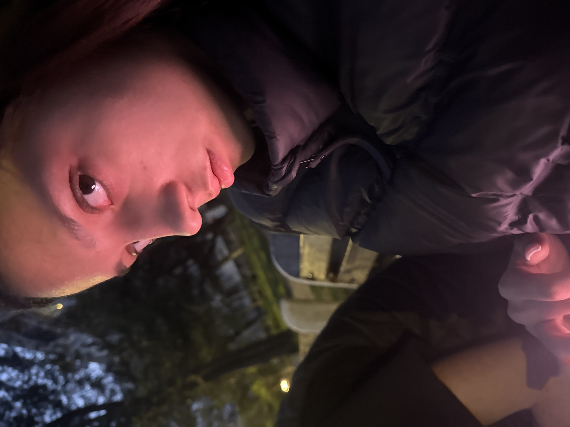The image is a close-up, rotated perspective of a person's face, illuminated by a red hue. The person is wearing a dark, puffy jacket. Part of a bench is visible in the mid-ground. The background is obscured, with hints of foliage and faint lighting. The location is not discernible based on the image alone.