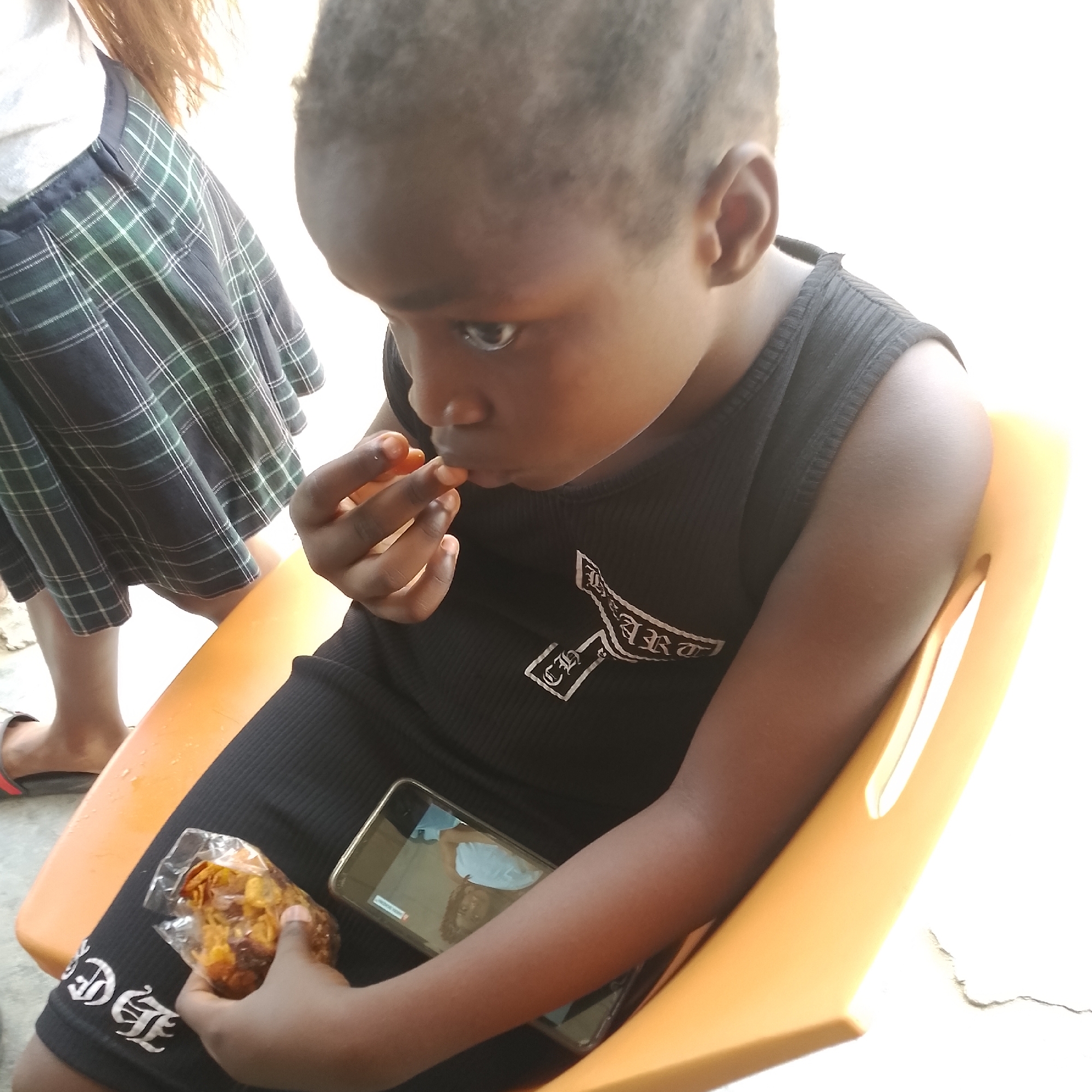 In the city of Ado, Nigeria, a young Black child is seated on an orange plastic chair. The child is wearing a black sleeveless top with a ribbed texture and a white graphic on the front. In their right hand, the child holds a clear plastic bag containing snacks, and their left hand is positioned near their mouth as if eating. A smartphone is visible on the child's lap, displaying an image. To the left of the child, another person, partially visible, wears a green and blue plaid skirt and a light-colored shirt. The background is bright and appears to be an outdoor setting, possibly a street or yard, with a white wall or building visible.