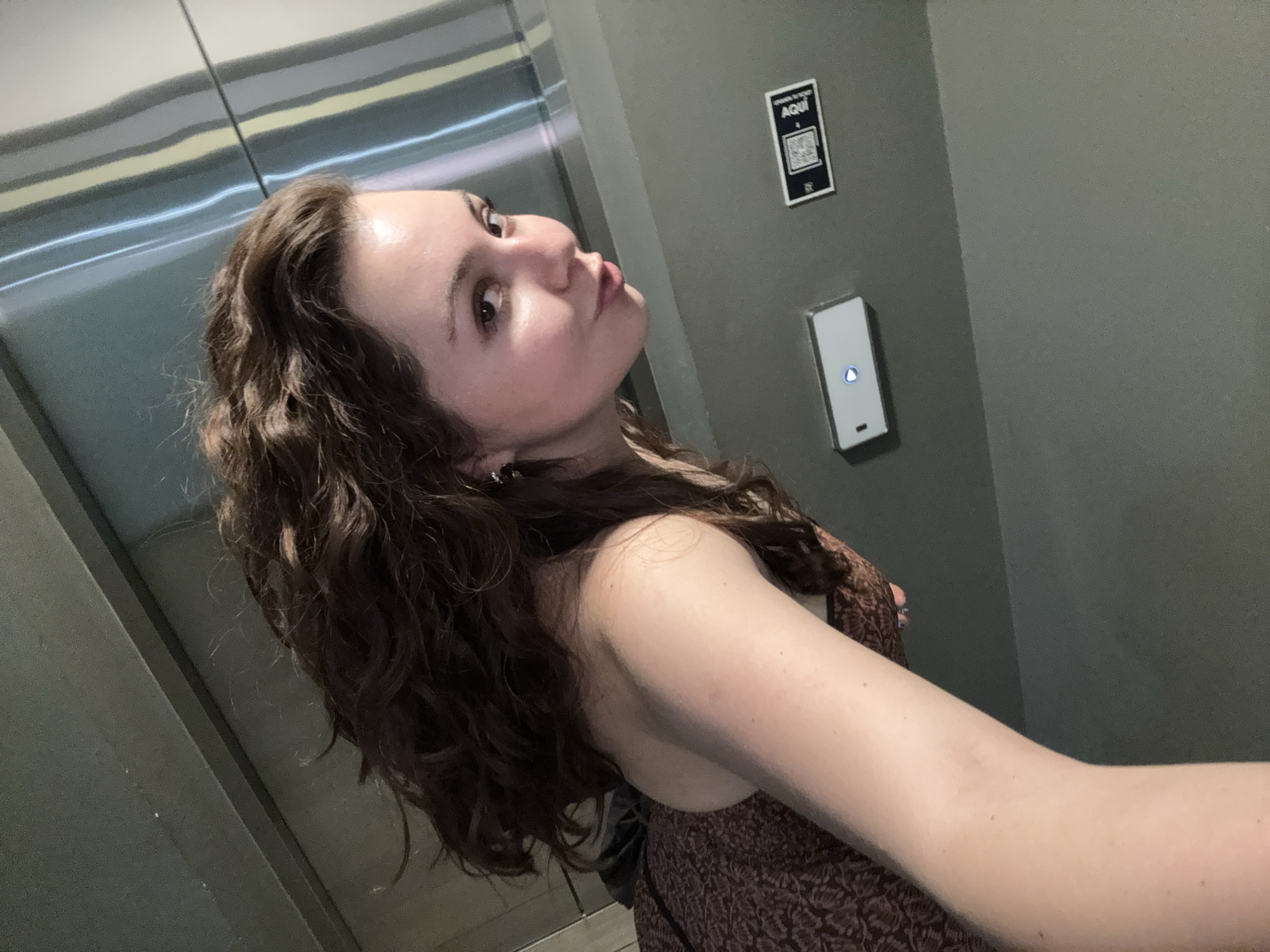 A young woman with curly brown hair is taking a selfie in what appears to be an elevator. She is wearing a brown patterned dress. The elevator has a metal door with horizontal lines and a grey wall with a small sign containing a QR code and the word "AQUI". Below the sign is a rectangular button with a blue light illuminated on it. The exact location in Mexico City is not discernible from the image.
