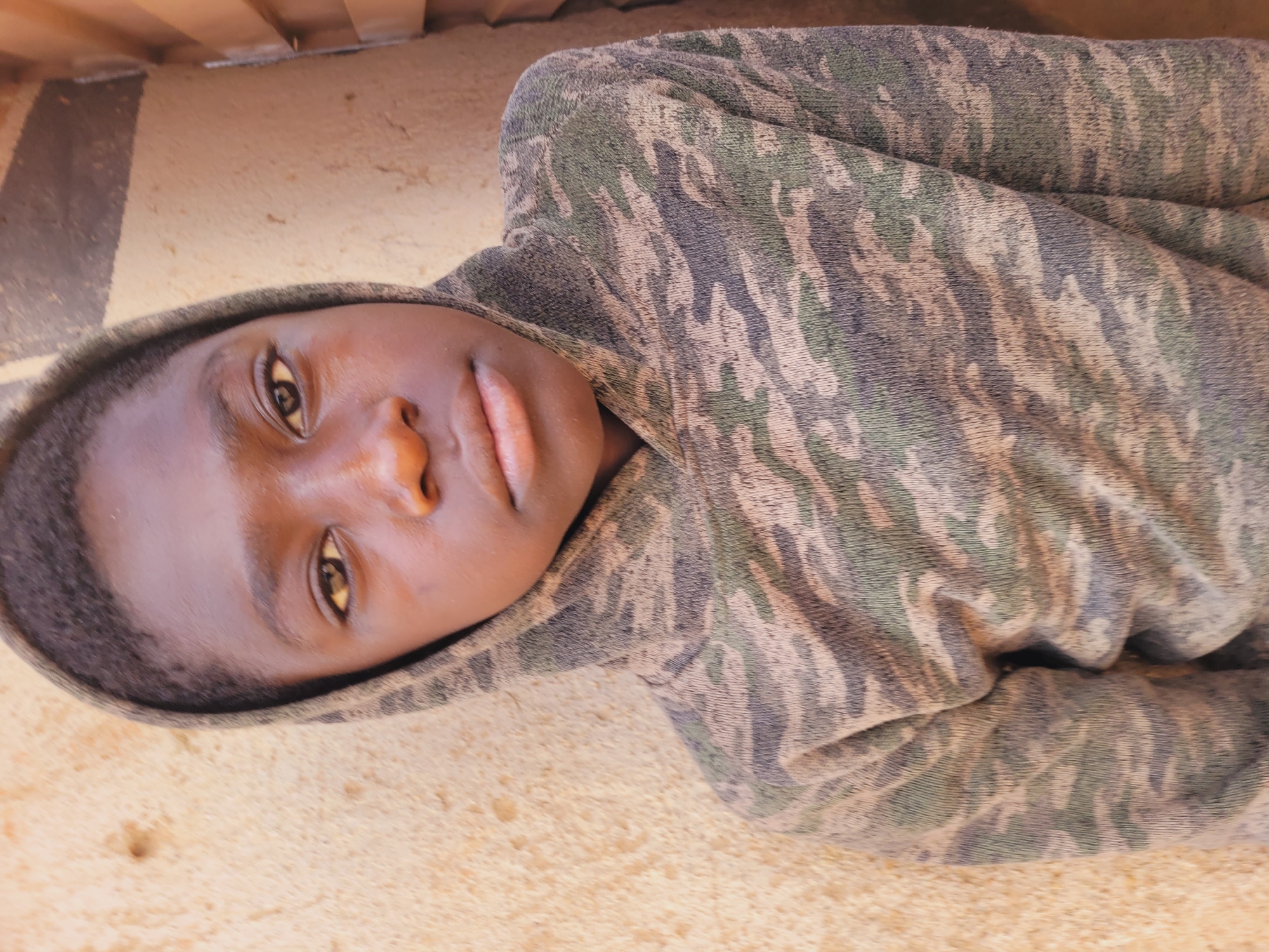 A young person with dark skin and brown eyes is lying on a tan surface, possibly the ground or a bed. The person is wearing a hooded sweatshirt with a camouflage pattern in shades of green, gray, and white. The hood is up, framing the person's face. The person's expression is neutral, with a slight upward gaze. The background is blurry and out of focus, but suggests an indoor setting with wooden elements. There is no evidence of Udu, Nigeria.