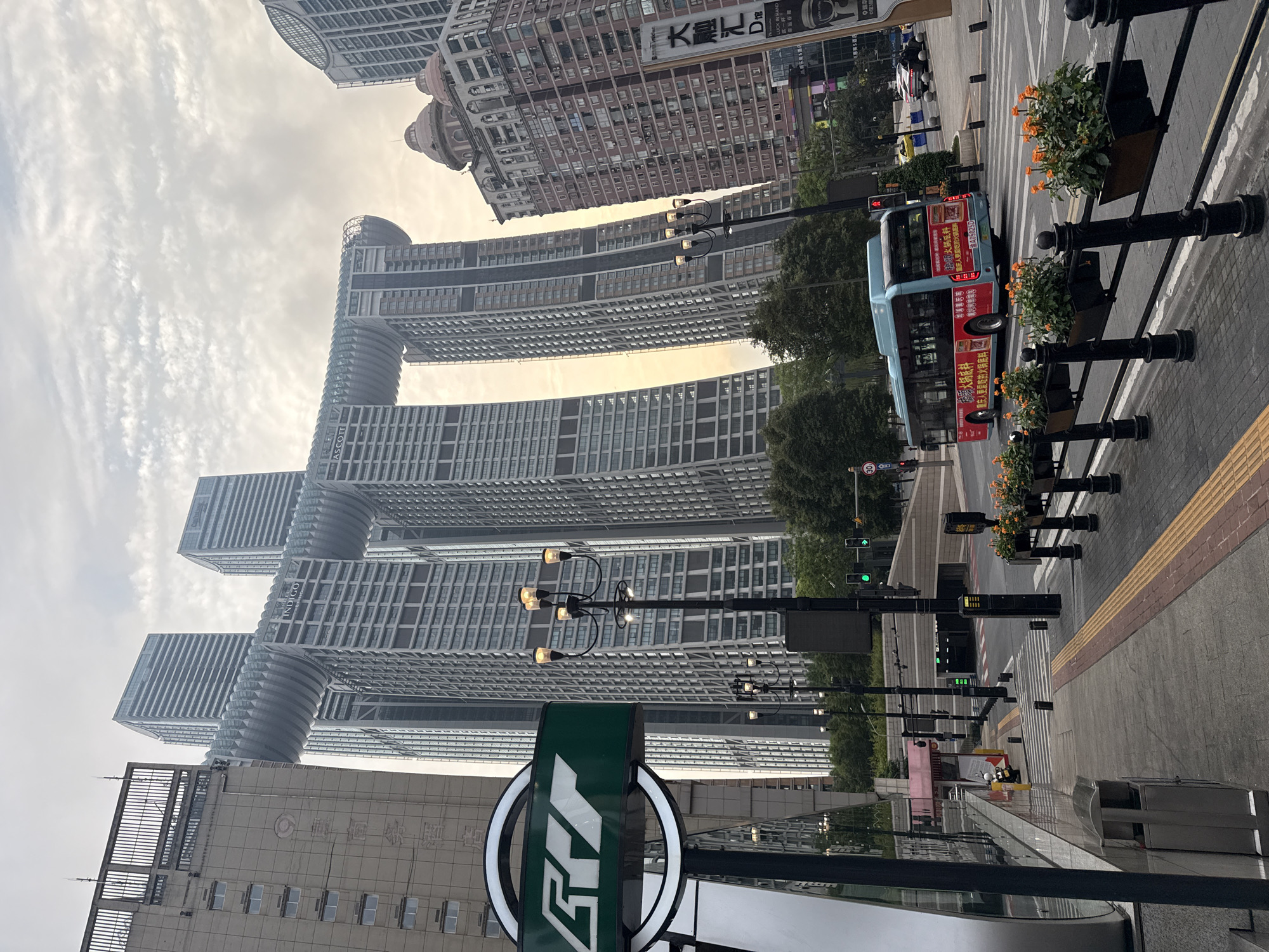The image captures a street scene in Yuzhong District, China, dominated by the distinctive Raffles City Chongqing architecture. The iconic twin towers, connected by a horizontal sky bridge, rise prominently against a cloudy sky. In the foreground, a green metro station sign with a stylized logo is visible on the left. A lamppost with multiple lights stands to the right of the sign. A light blue bus with red advertisements is driving across the intersection. Traffic lights, pedestrian crossing, and street planters with orange flowers are also present. The overall environment suggests a bustling urban area with modern infrastructure.