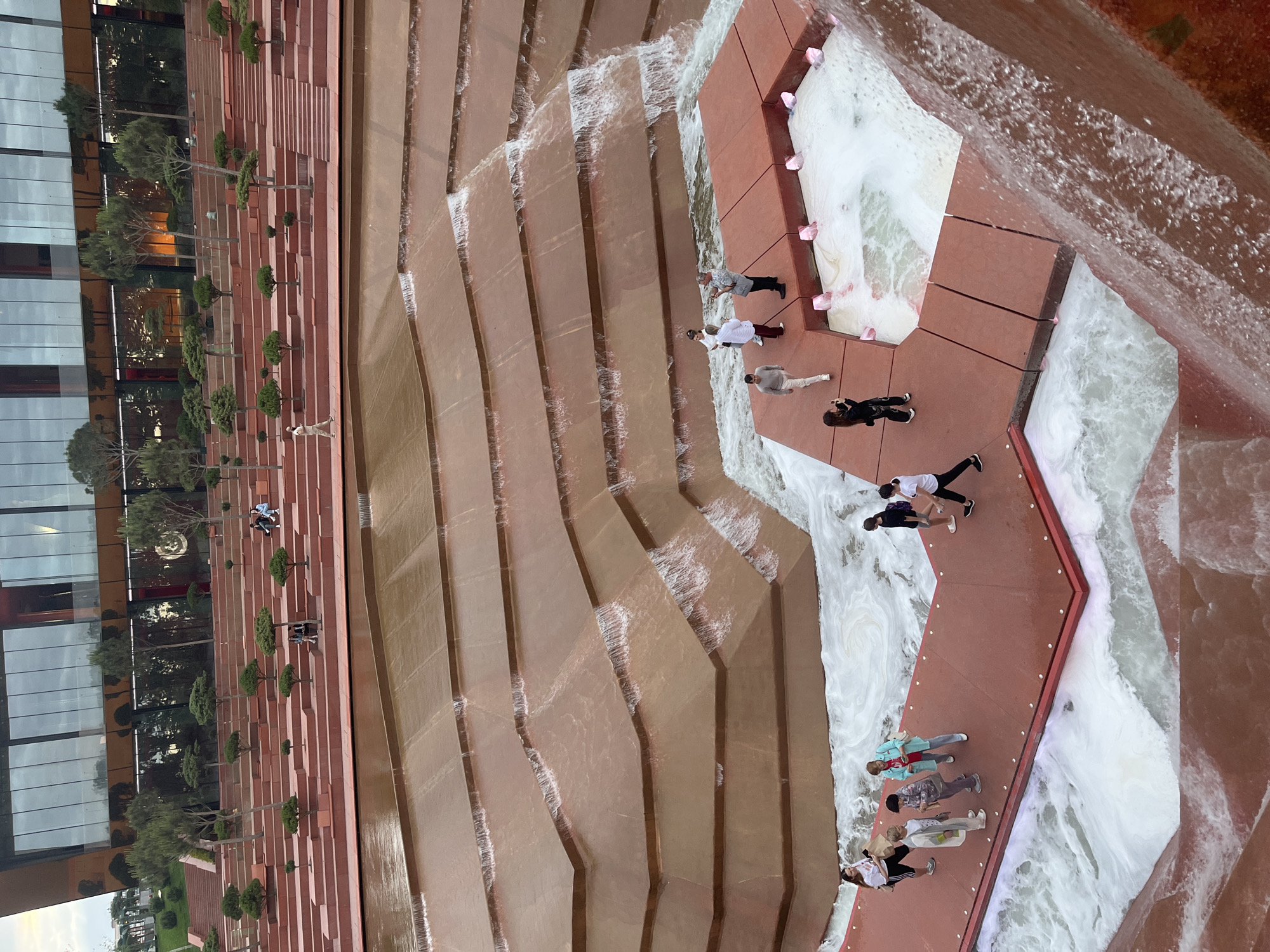 The image shows an elevated view of the Krasnodar Fountain in Krasnodar, Russia. The fountain is constructed as a series of stepped platforms that create a cascade effect for the water. Several people are walking along the red walkways bordering the flowing water. A modern glass building and rows of small trees are on the left side of the fountain. The water is moving down the stepped tiers, creating white foam where it falls. The environment suggests an urban park setting.