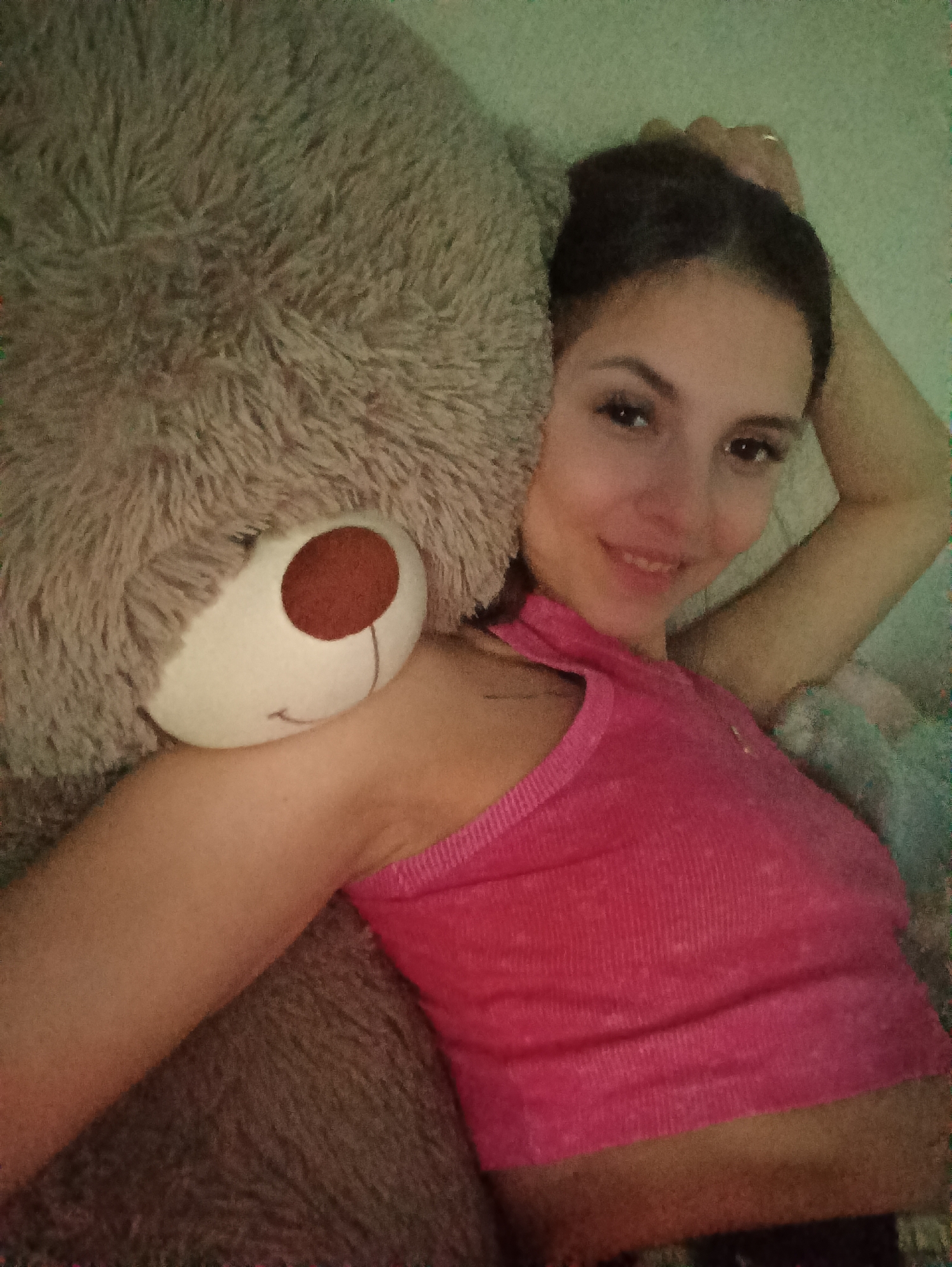A young woman is shown lying comfortably next to a large, fluffy teddy bear in what appears to be an indoor setting, likely a bedroom or lounge area. The woman occupies the right side of the frame, smiling gently at the camera. She has dark hair, styled away from her face, and dark eyes, with subtle makeup highlighting her eyes. She is wearing a bright pink ribbed tank top. Her right arm is raised, with her hand resting behind her head, revealing a ring on her ring finger.
The teddy bear, a dominant subject on the left, is light brown or beige with a soft, shaggy texture. It features a distinctive white snout with a large brown nose and a subtle stitched smile. The woman's left arm is draped over the bear, suggesting a close and comfortable interaction.
The background consists of soft, light-colored surfaces, possibly bedding or cushions, creating a cozy and relaxed atmosphere. The lighting is soft and warm, enhancing the intimate feel of the scene. The woman's relaxed posture and pleasant smile convey feelings of contentment and warmth. There is no visible text in the image.