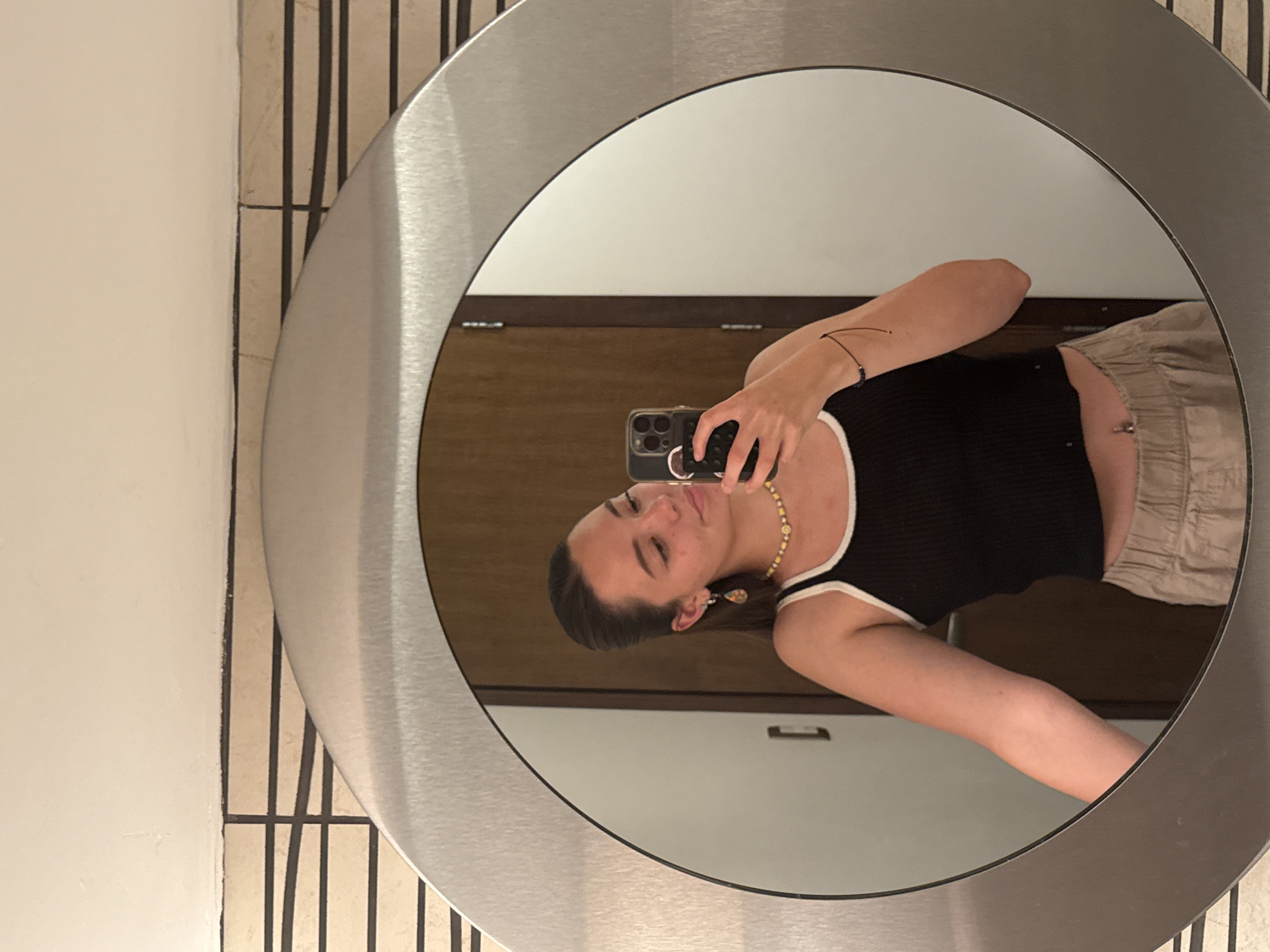 A person is taking a selfie in a circular mirror in a bathroom. The person is wearing a black tank top with white trim and tan pants. They are holding a phone in their right hand. The background includes a wooden door with a handle, a light beige wall with vertical black and tan stripes, and a white wall. The location is likely a private bathroom, but there are no elements to confirm the exact location of San Bernardino Tlaxcalancingo, Mexico.