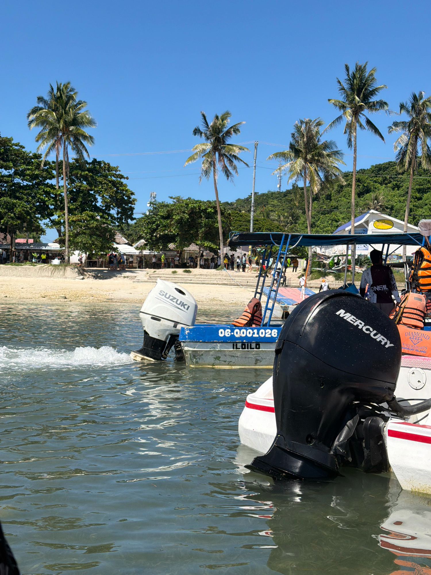 This image depicts a sunny day on the waters of Boracay Island in Malay, Philippines, likely near White Beach. In the foreground, a white boat with a prominent black Mercury outboard motor is partially visible. The motor is submerged in the clear, choppy water, leaving a trail of white spray. Another boat, painted blue and white, is positioned behind the foreground boat, also equipped with an outboard motor, this one bearing the "SUZUKI" logo. This second boat has a canopy structure providing shade and is adorned with orange life vests. In the background, a sandy beach is lined with palm trees and lush green vegetation. Several structures, possibly shops or establishments, are visible along the beachfront. A few people are scattered on the beach and near the boats, engaged in various activities, suggesting a tourist destination. The sky is a vibrant, cloudless blue.