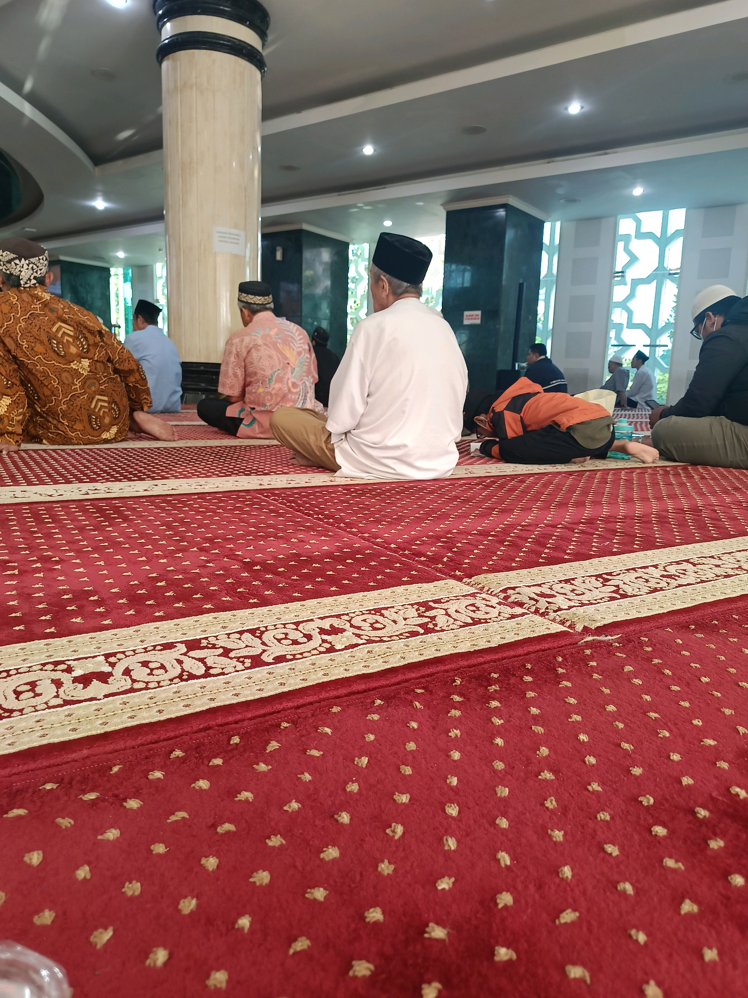 The image shows a group of men sitting and praying on a red carpet in what appears to be a mosque. In the foreground, the red carpet has a repeating pattern of small, light-colored dots. A decorative border runs across the carpet, featuring intricate beige and red patterns. Several men are visible, seated or in prayer postures.
On the left, a man in a brown patterned robe and a black cap is seen from the back. To his right, another man in a pink patterned shirt and black cap is also seated. In the center, a man in a white long-sleeved shirt and black cap is in a prayer posture, with his head bowed to the ground. To his right, a younger person in an orange jacket and black pants is also in a prayer posture. Further back, other individuals are visible, including a man in a black jacket and white cap on the far right.
The environment includes a large, fluted pillar with a black band at the top, located to the left of center. The ceiling is visible, with recessed lighting. In the background, there are windows with a distinctive geometric pattern and what appears to be a dark green wall. The overall impression is a serene and communal prayer setting.
The location is identified as a mosque in Banguntapan, Indonesia. This detail provides specific geographical context for the scene depicted.