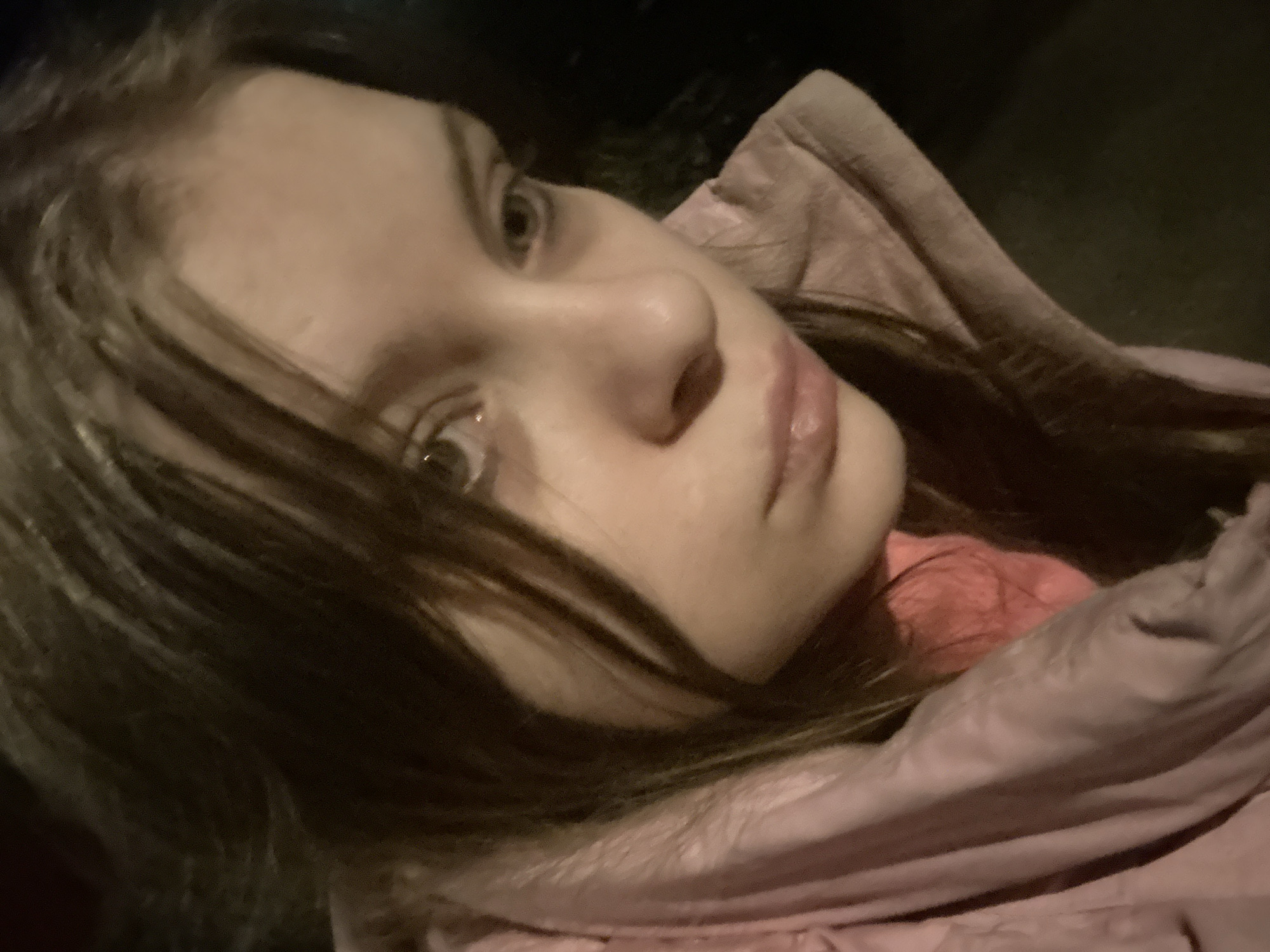 A close-up, low-angle selfie captures a young woman with fair skin and light brown hair. She is wearing a pink puffy jacket with the collar turned up. Her eyes are light-colored, and she has a neutral expression. The background is dark and out of focus, likely an urban setting at night. The location is not discernible as Moscow, Russia based on the provided image.