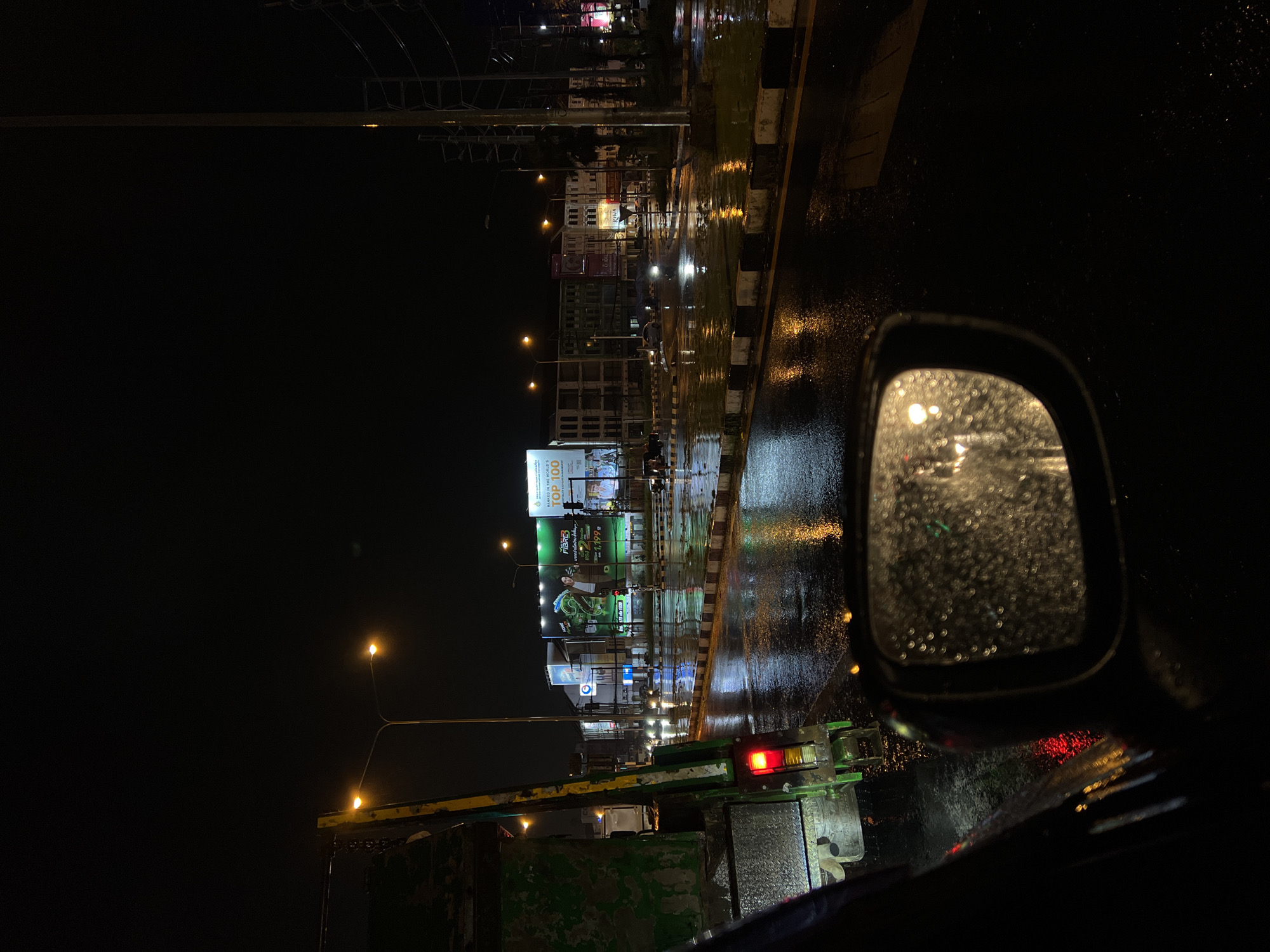 Captured from within a vehicle, the scene unfolds in the city of Unknown on a rainy night. The driver's side mirror, speckled with raindrops, reflects blurry lights. The street outside is wet, reflecting the glow of streetlights and illuminated billboards. A green garbage truck is partially visible. No people are seen.