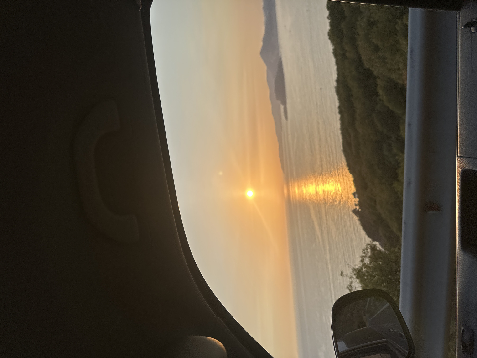 The image depicts a view from inside a vehicle, looking out the window at a scenic sunset over the sea. The sun is low on the horizon, casting a golden reflection on the water. In the distance, a mountainous coastline is visible, possibly the area around Sveti Stefan in Montenegro, given the characteristic coastal landscape. The foreground shows a portion of the car's interior, including the door panel and a rearview mirror, which reflects a winding road. The overall atmosphere is tranquil and serene, capturing a moment of natural beauty during a road trip.