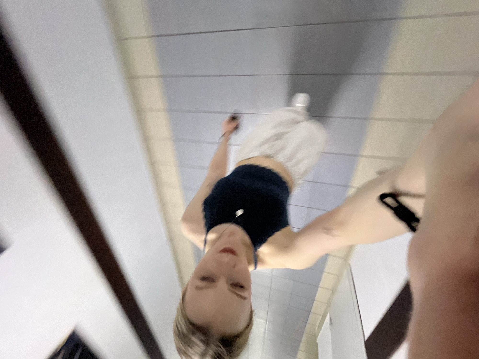 The image shows a woman taking a selfie in what appears to be a public restroom. She is wearing a navy blue tank top and white shorts. She is looking at the camera with a neutral expression. Her hair is blonde and appears to be in a short hairstyle. The background suggests a tiled room, possibly a bathroom. The specific location within Khimki, Russia, is not discernible from the image alone.
