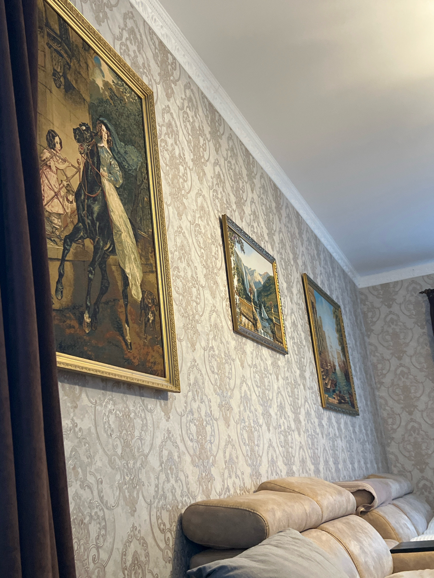 This image captures an interior scene of a room decorated with ornate wallpaper featuring a damask pattern. Three framed paintings are displayed on the wall. The largest painting, on the left, depicts a historical scene with a woman on horseback, attended by a figure in pink. A dark brown curtain is partially visible on the left side of the frame. To the right of the large painting, a smaller landscape painting shows a waterfall and a sailboat. Further to the right, another painting displays a cityscape with a body of water. In the foreground, a beige sectional sofa is visible, partially obscuring the bottom of the wall and paintings. The ceiling is white and features decorative molding along the wall-ceiling junction. The scene appears to be a living room or a similar domestic space, potentially located in Levino, Russia. There are no people actively engaged in actions.