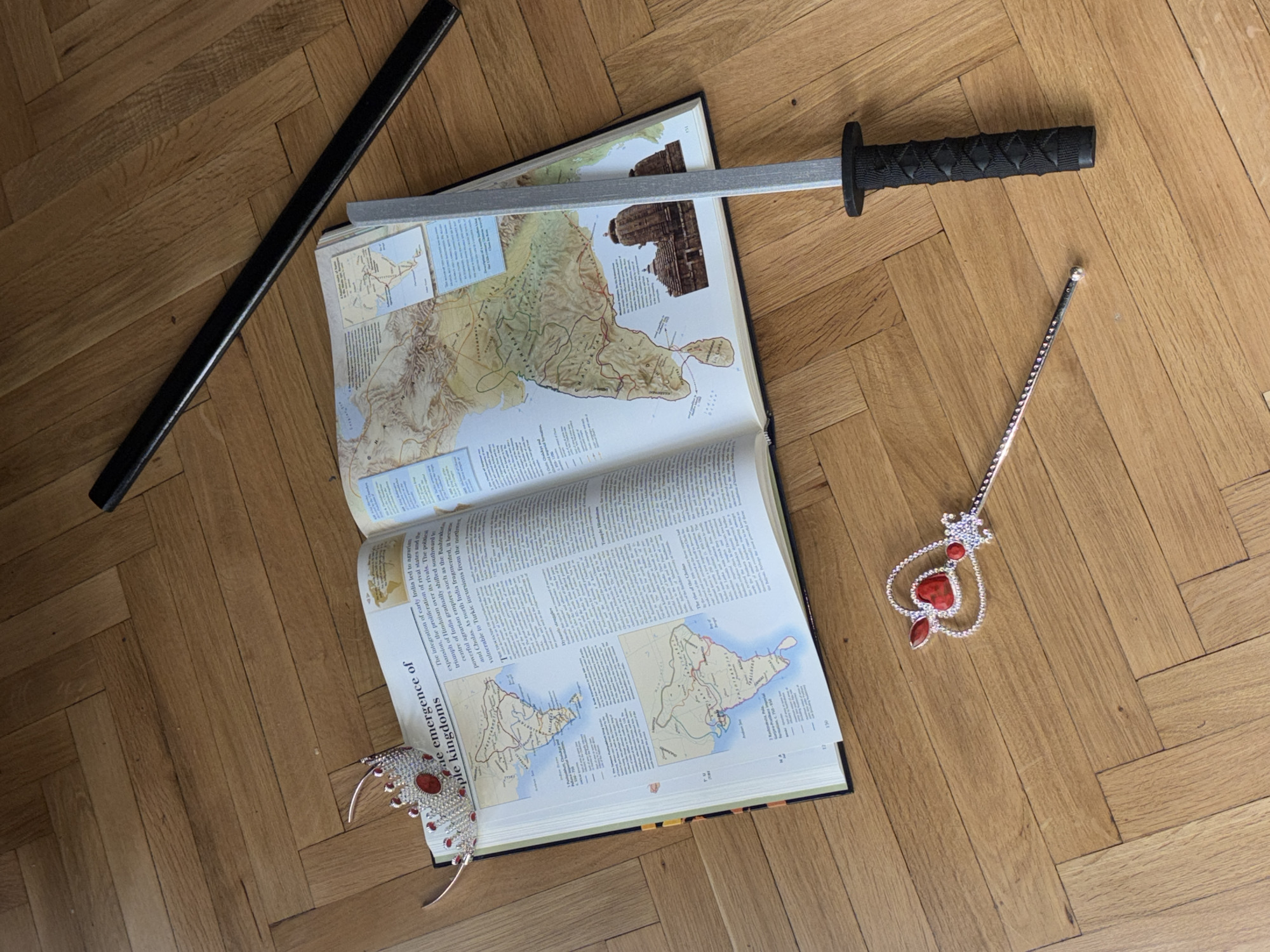 The scene depicts an open book lying on a wooden floor. The book appears to be a textbook, featuring maps and text about the emergence of kingdoms in India. On top of the book lies a toy sword with a black handle and a gray blade. To the left of the book is a black bo staff. To the right of the book is a toy scepter with red jewels. The floor is made of wood planks. The location (Varna, Bulgaria) is not discernible from the image.