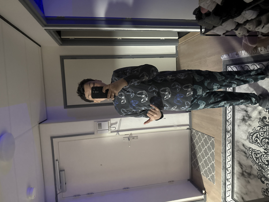 An individual is depicted in a full-body selfie taken in a mirror within an indoor residential setting, likely an apartment hallway or entryway in Voorburg, Netherlands. The subject, with short, dark, curly hair, holds a dark-colored smartphone to obscure their face. They wear a dark-colored, two-piece pajama or loungewear set featuring an all-over pattern of video game controllers in white and blue accents, along with dark grey or black slip-on slippers. A ring is present on the subject's left ring finger. Their right hand (viewer's left) performs a "shaka" or "hang loose" gesture.
The background reflected in the mirror reveals a white interior door with a dark frame, equipped with a silver-colored handle, a peephole, and an overhead hydraulic door closer. Adjacent to this door, a white intercom or entry system panel is mounted on the wall. The flooring consists of light-colored wood or laminate, with a smaller, grey and white patterned rug placed in front of the white door. A larger, dark patterned rug with ornate designs is visible in the foreground reflection. To the subject's right, another doorway with a dark frame leads into an unlit or darker interior space. On the far right, a dark metal multi-tiered shoe rack holds several pairs of shoes. The ceiling is white with a round, flush-mounted light fixture, and blue ambient light reflections are visible on the ceiling and walls.
