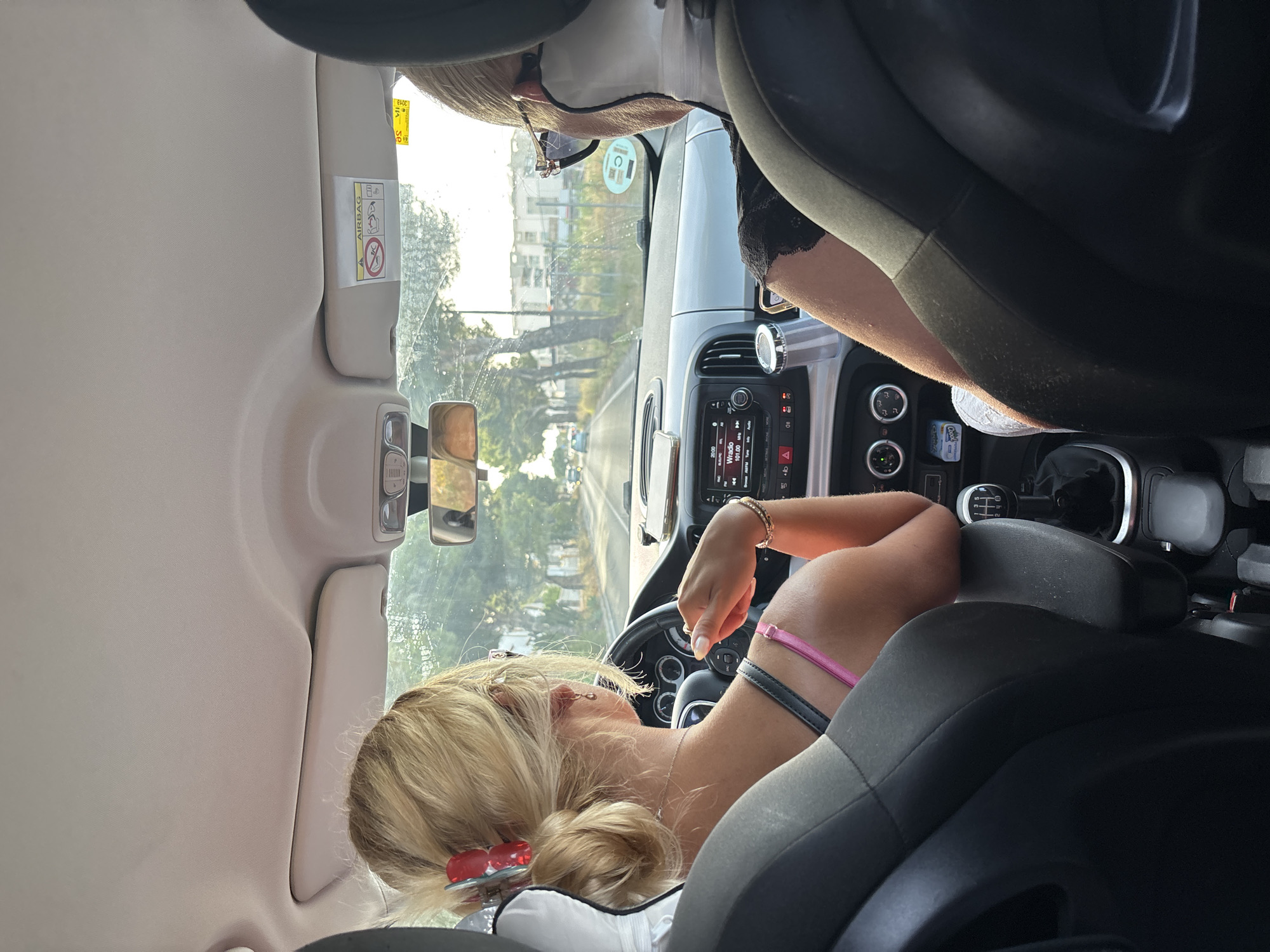 The image shows the interior of a car with two women inside, viewed from the back seats. The woman in the driver's seat has blonde hair pulled back with a red clip and is wearing a pink strap top and a necklace. The woman in the passenger seat is wearing sunglasses and a black top with lace detail. Both women appear to be in motion, as indicated by the road visible through the windshield and the slight blur of the trees outside. The car's interior is visible, including the dashboard with a radio display, gear shift, and various buttons and vents. The image was taken during the daytime, with sunlight illuminating the scene. The location is identified as Orihuela, Spain, though no specific landmarks are clearly identifiable to confirm this within the image itself.