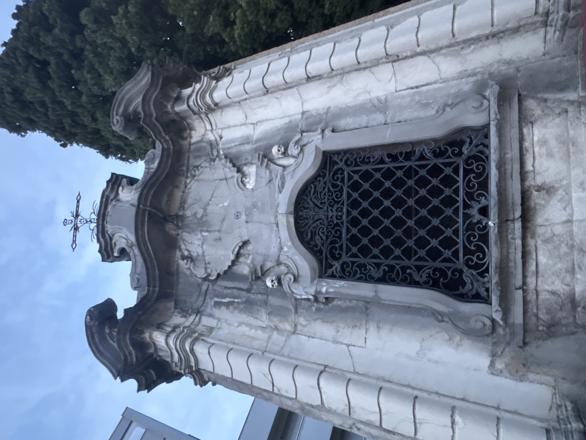 The image shows a detailed view of a stone mausoleum in Balerna, Switzerland, against a backdrop of a blue sky and a tall evergreen tree. The mausoleum features ornate architectural elements, including decorative carvings and a wrought-iron grate covering a window. Two small skull carvings are visible on either side of the window. The structure appears to be old, with signs of weathering on the stone. A modern building is partially visible on the left side of the frame. The lighting suggests it might be early morning or late evening.