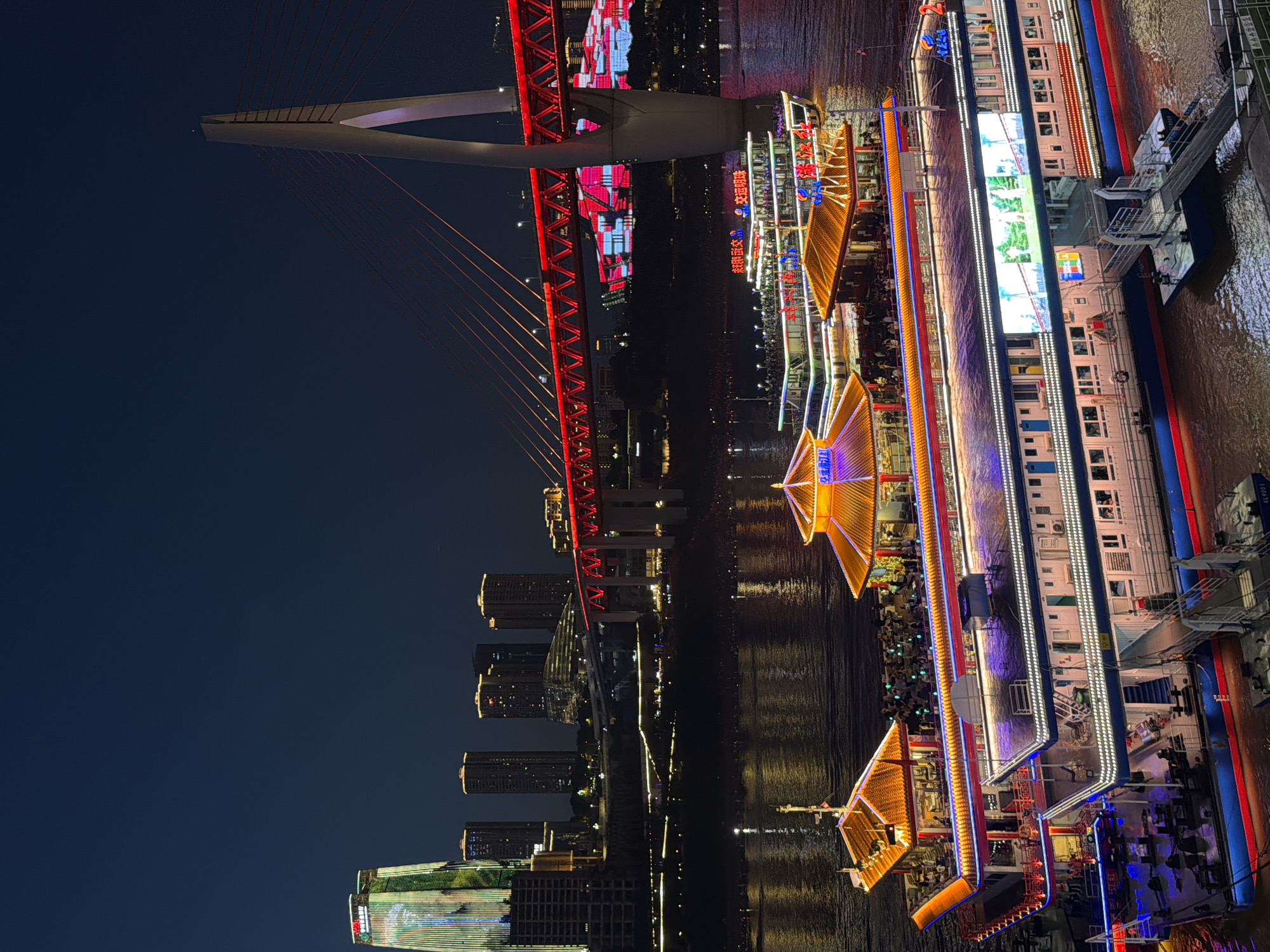 This is a nighttime photograph of a city skyline and a river with a large boat docked on the right. The scene is illuminated by a vibrant display of lights from the cityscape, the bridge, and the boat.
The main subjects are the Hongyadong Bridge, a suspension bridge with a red truss structure and white towers, and a multi-story passenger boat docked on the river. The bridge is adorned with red and pink lights, with cables stretching from the towers. The boat is brightly lit with strings of white and purple lights along its length, and has illuminated sections with orange triangular roofs. The river reflects the lights, creating a shimmering effect. In the background, several tall buildings with illuminated windows form the city skyline.
There are people visible on the deck of the boat, suggesting it is in operation or at least accessible. The overall atmosphere is one of a lively urban nightscape. The time of day is clearly night, with a dark sky. The weather appears calm, with no indication of rain or strong winds.
The location is hinted to be Yuzhong District, China. Some Chinese text is visible on the boat, and on the buildings in the background, but it is too small to be legible.
The image captures a dynamic and colorful urban scene, emphasizing the city's nightlife and its connection to the river.